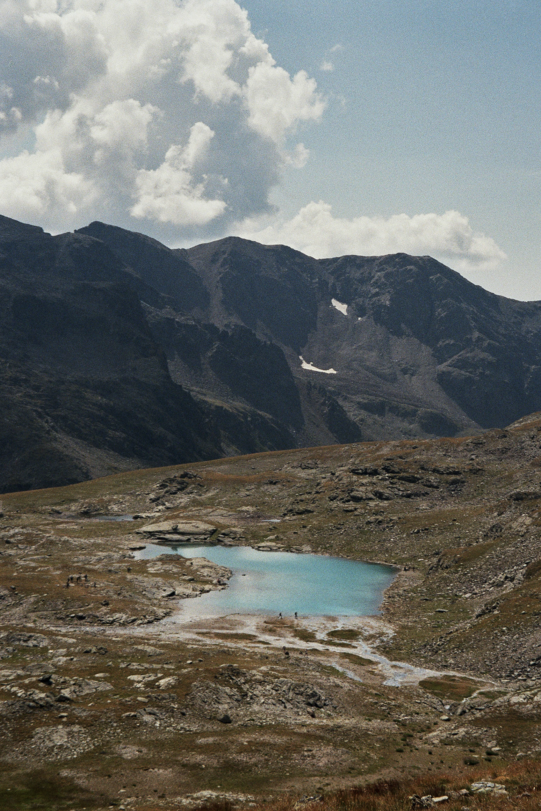 Mountain landscape with cloudy sky, rugged peaks, small lake, and hikers in the foreground.