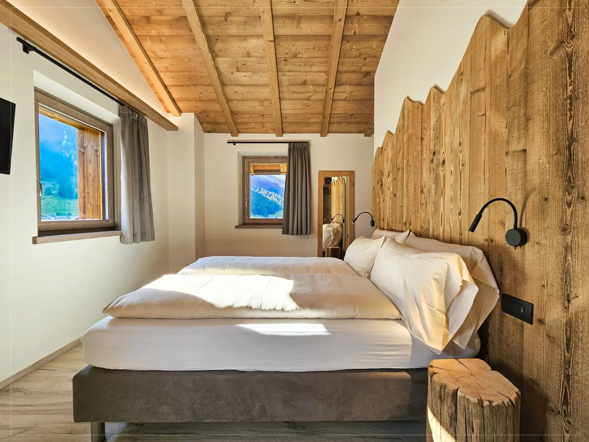 The image shows a cozy bedroom with two beds, a large window with curtains, wooden ceiling with beams, and a wooden headboard with built-in reading lights. A mirror and small wooden stool are also visible.