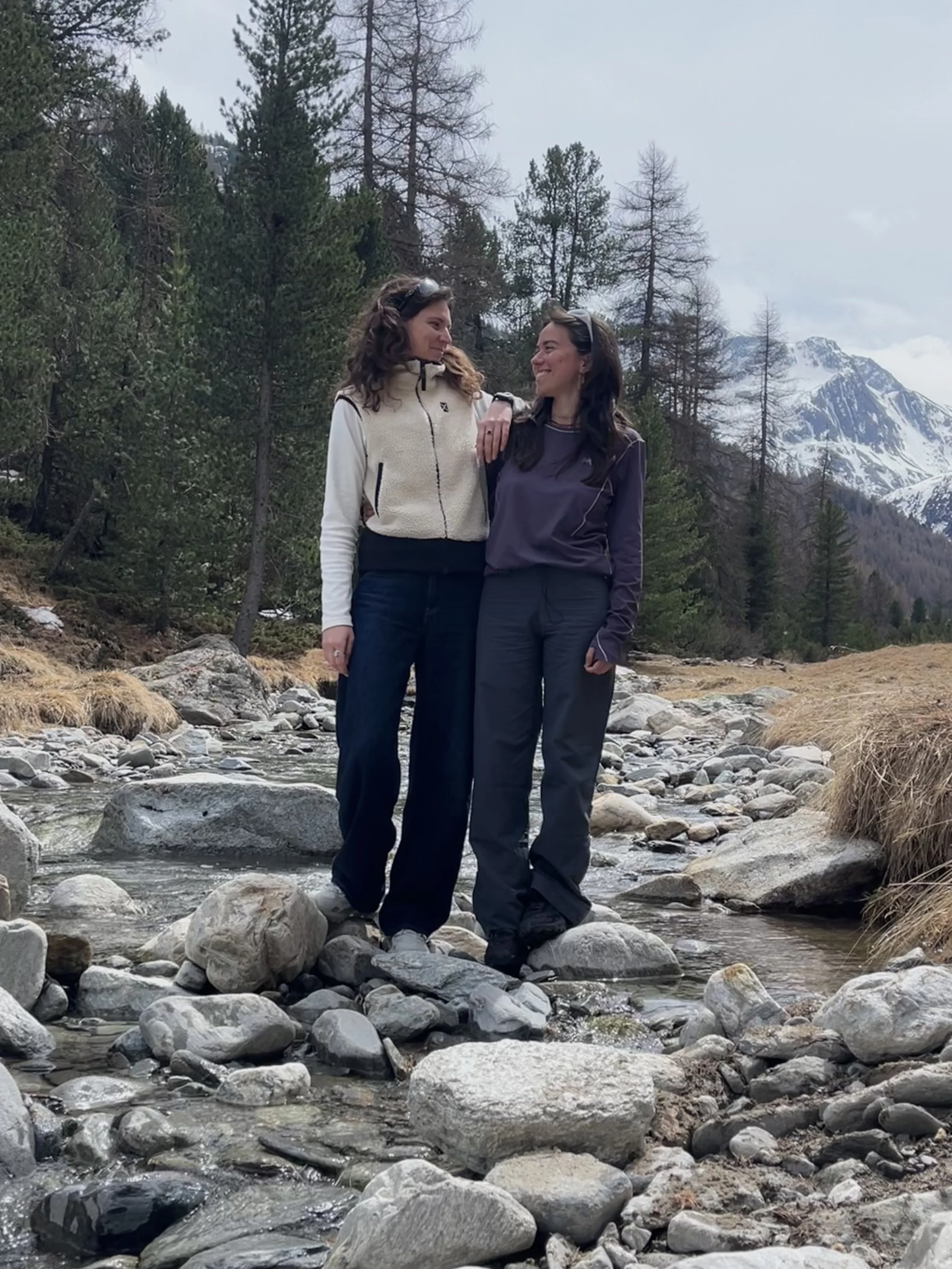 Two women stand close together on a rocky stream bed in a mountainous, forested landscape with snow-capped peaks in the background. One woman has her arm around the other, and both are smiling.