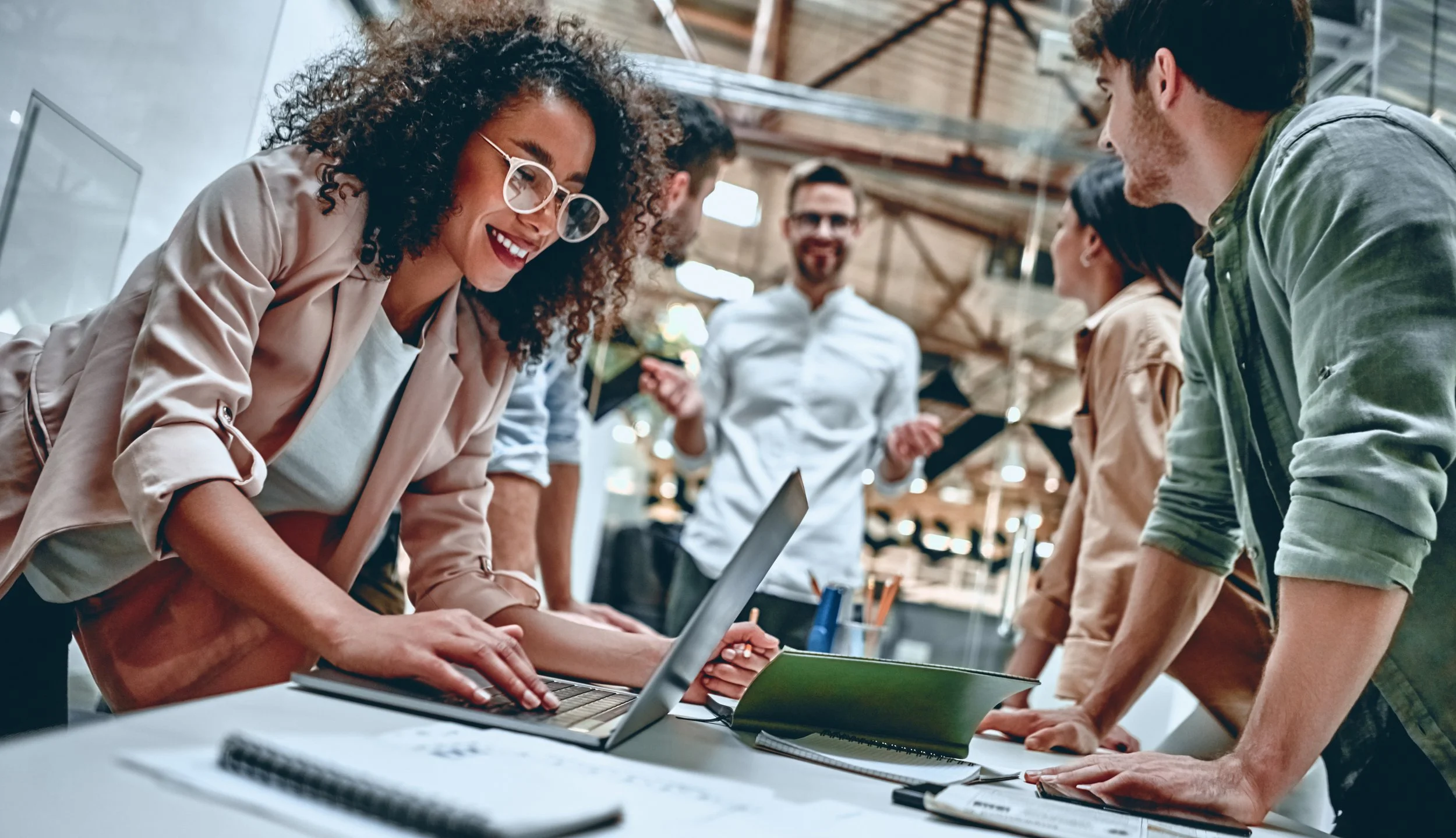 A group of diverse young professionals collaborating around a table in a modern, open office space. One woman with curly hair and glasses is smiling as she works on her laptop, while others engage in discussion and review documents.