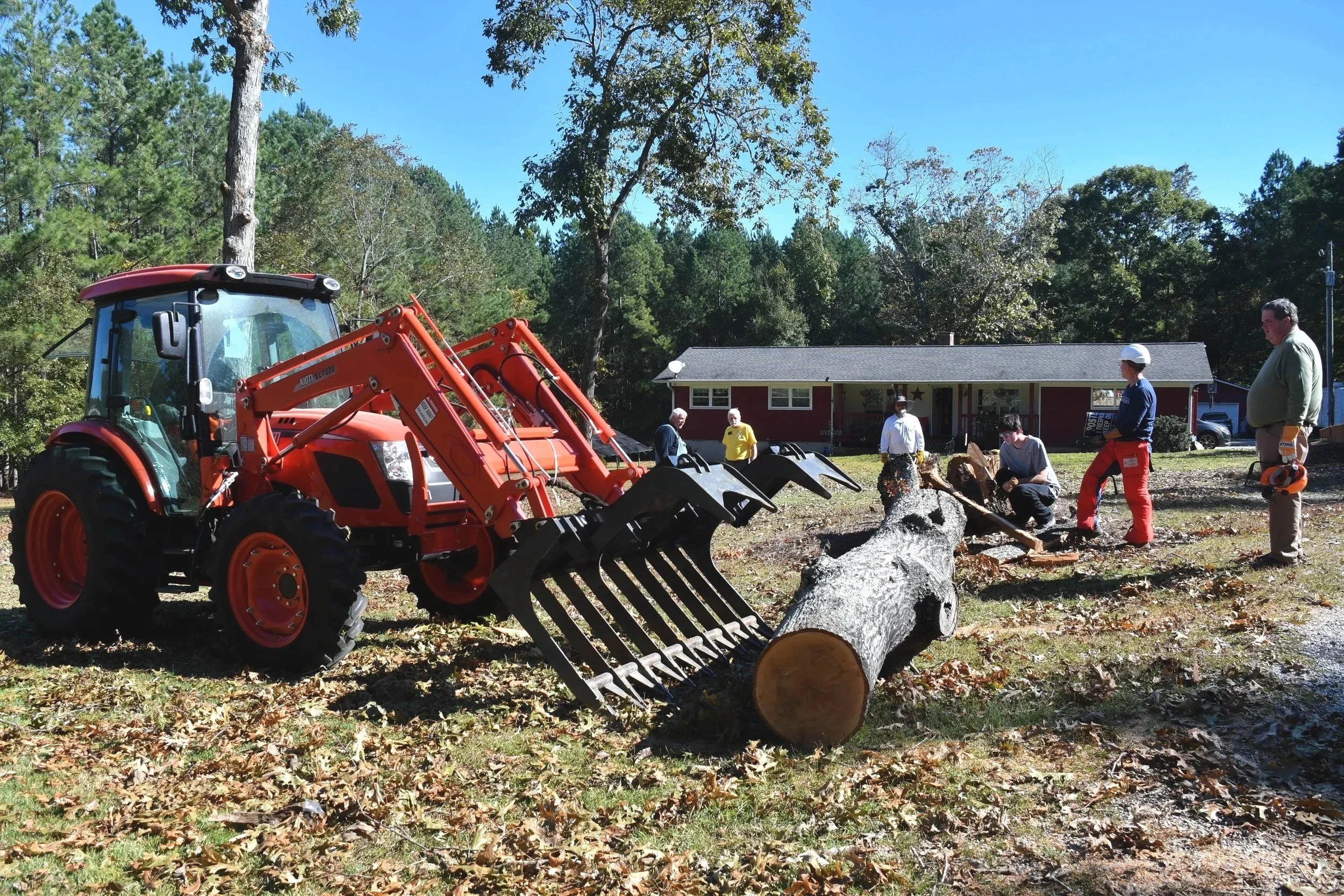A group of people gathered outdoors in front of a house, with a red tractor and a large fallen tree. The scene appears to be a community event or tree removal activity on a clear sunny day.