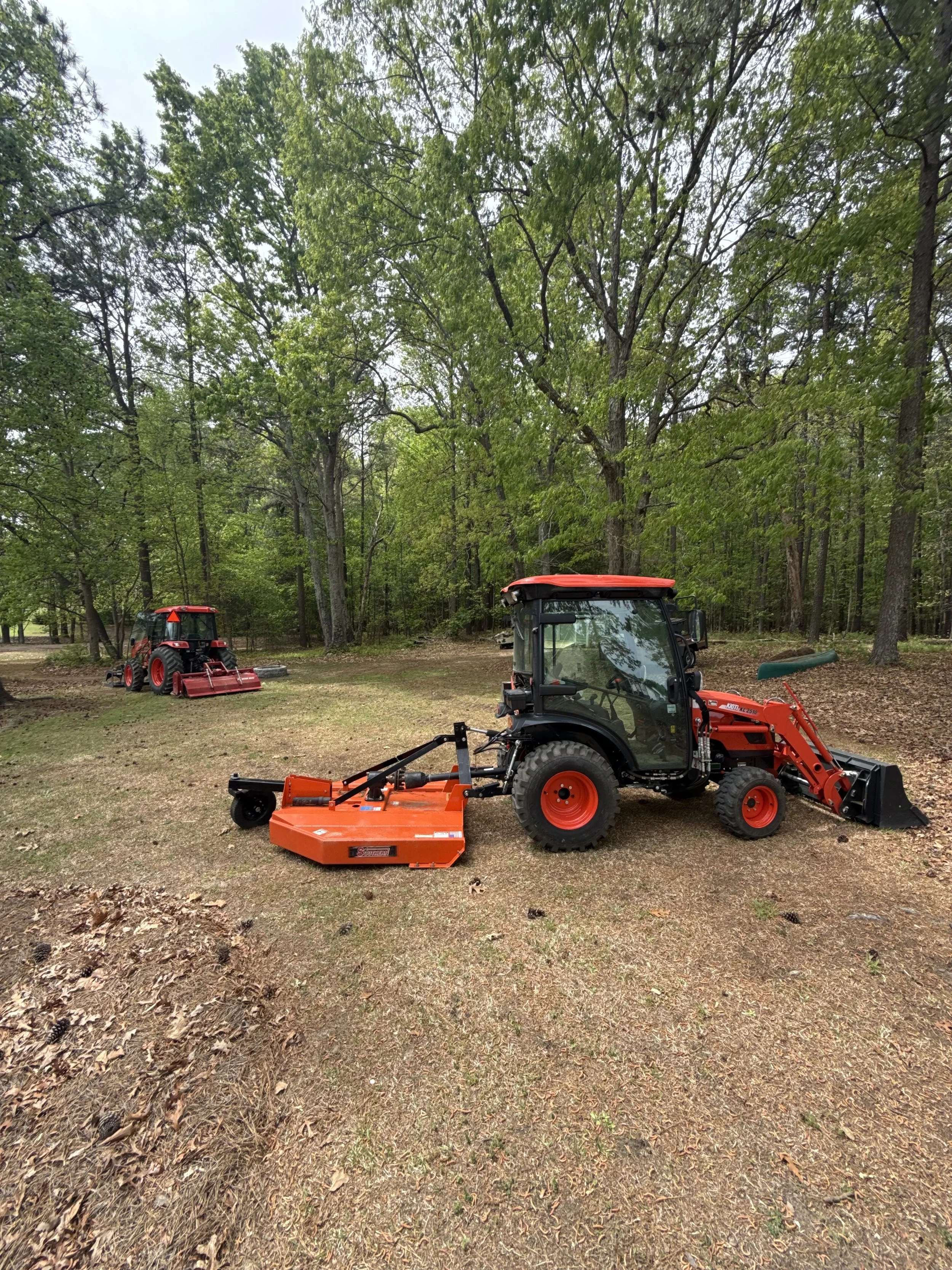 A lawn tractor with an attached mower deck parked on a grass field surrounded by trees in a wooded area.