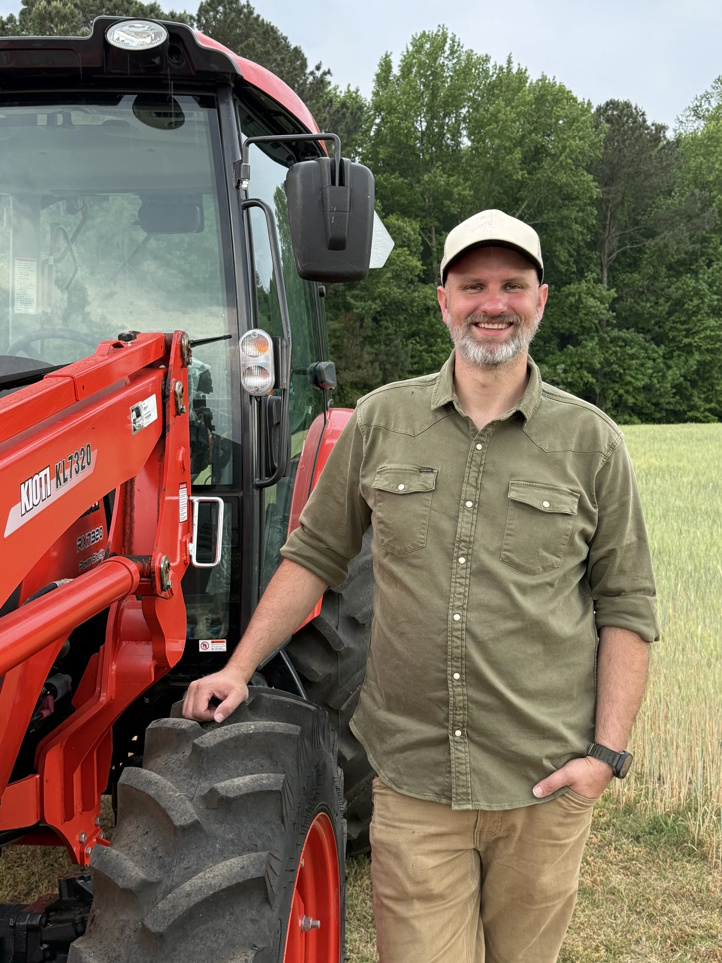 A smiling man in a khaki shirt and beige cap standing next to a red tractor in a grassy field with trees in the background. Gary with one of his trusty tractors.
