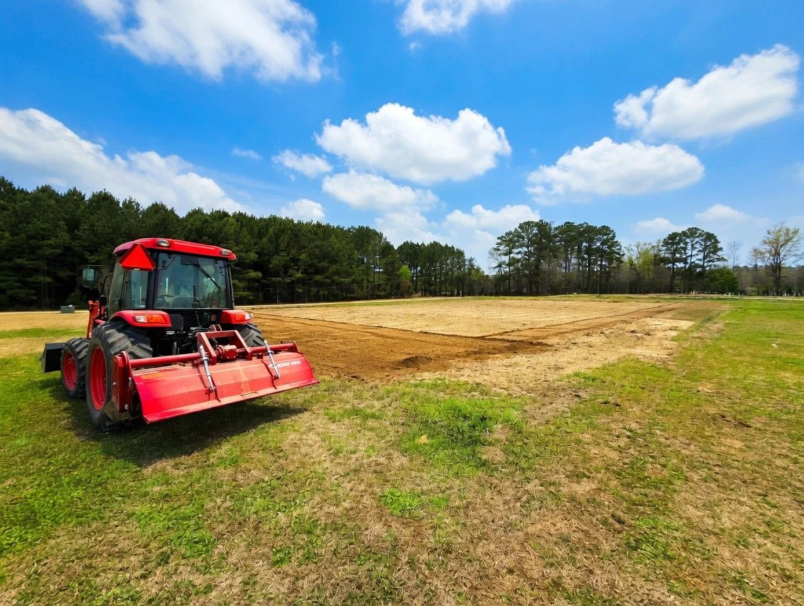 A red tractor on a grassy field with a patch of tilled dirt, under a partly cloudy sky with trees in the background.