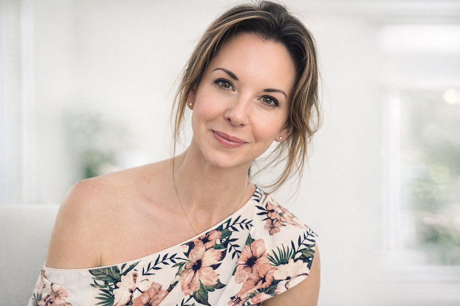 Portrait of a woman with shoulder-length hair, smiling, wearing a floral off-shoulder top, earrings, and a necklace, in a bright room with white walls and a window.