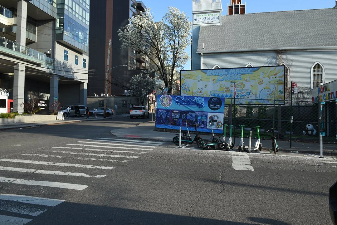 Street scene with electric scooters parked on the sidewalk, a crosswalk in the foreground, and a colorful billboard advertising a magic castle attraction in the background.