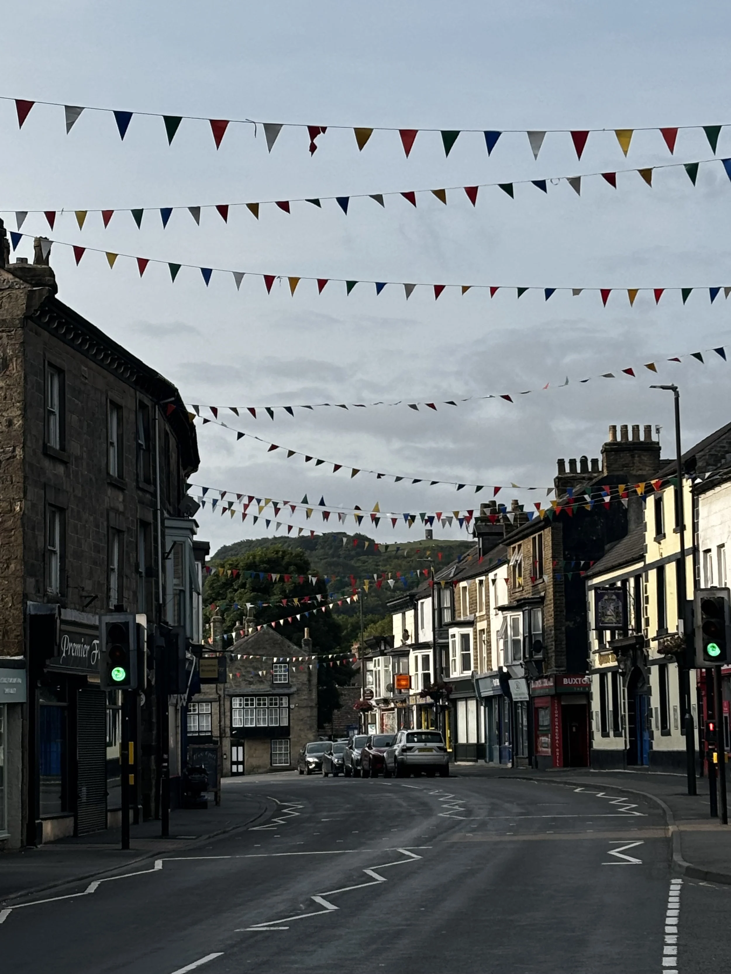 Buxton street decorated with colourful triangular bunting flags hanging across the road between historic buildings and shops on a bright, overcast day.