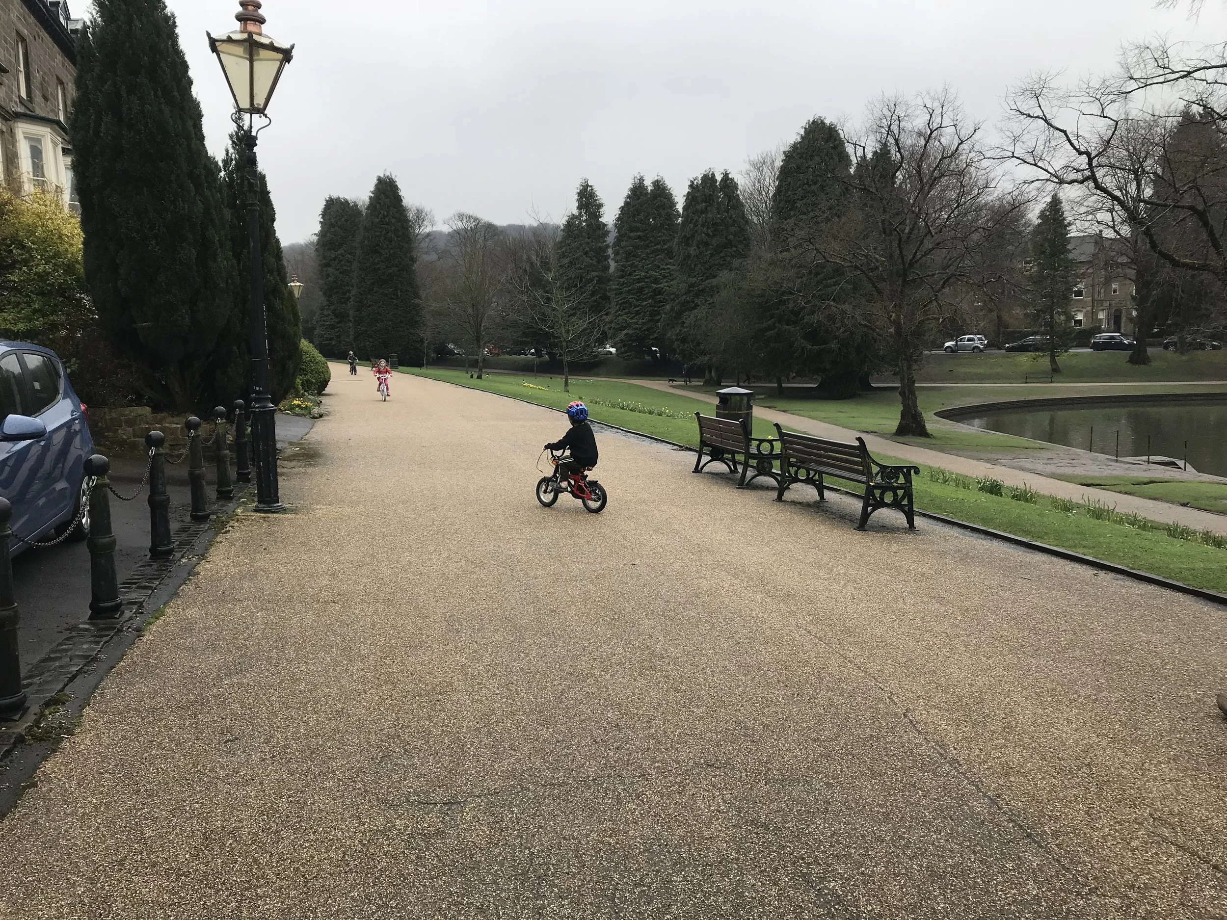 A young child riding a bicycle along a paved pathway in Buxton Pavilion Gardens, with park benches, the pond, and mature trees in view