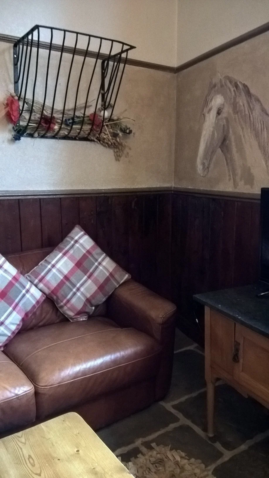 A rustic corner of the living room featuring a brown leather sofa with plaid cushions, traditional wooden wall paneling, and an wall painting of a horse at The Old Stables Buxton.