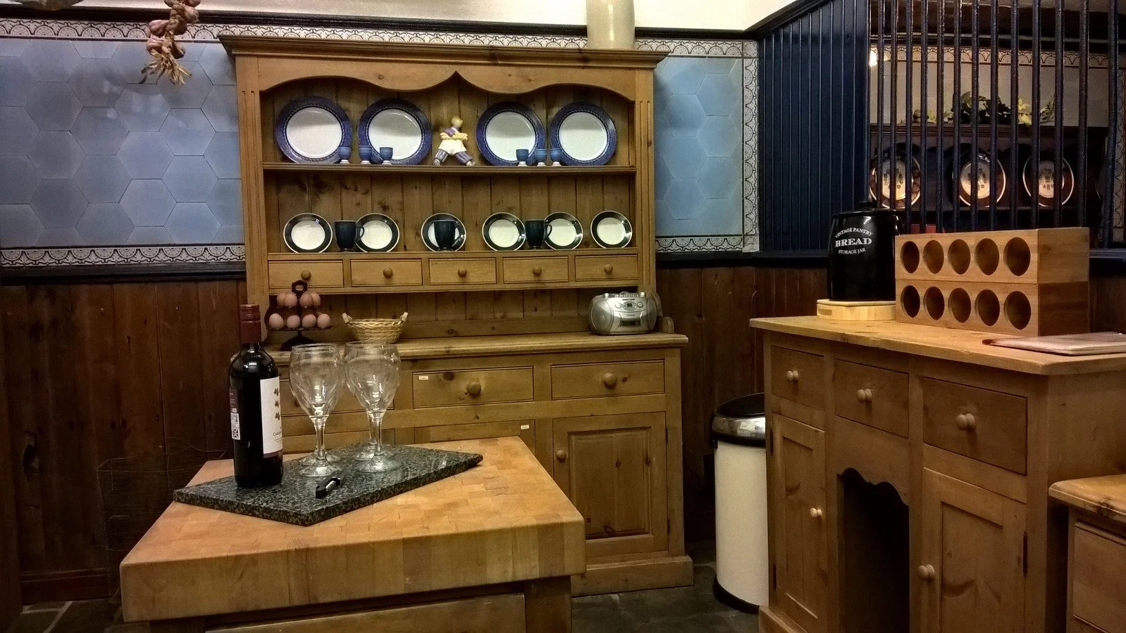 Kitchen with wooden cabinets, shelves displaying plates and cups, a table with wine bottle and glasses, and various kitchen decor at the Old Stables Buxton
