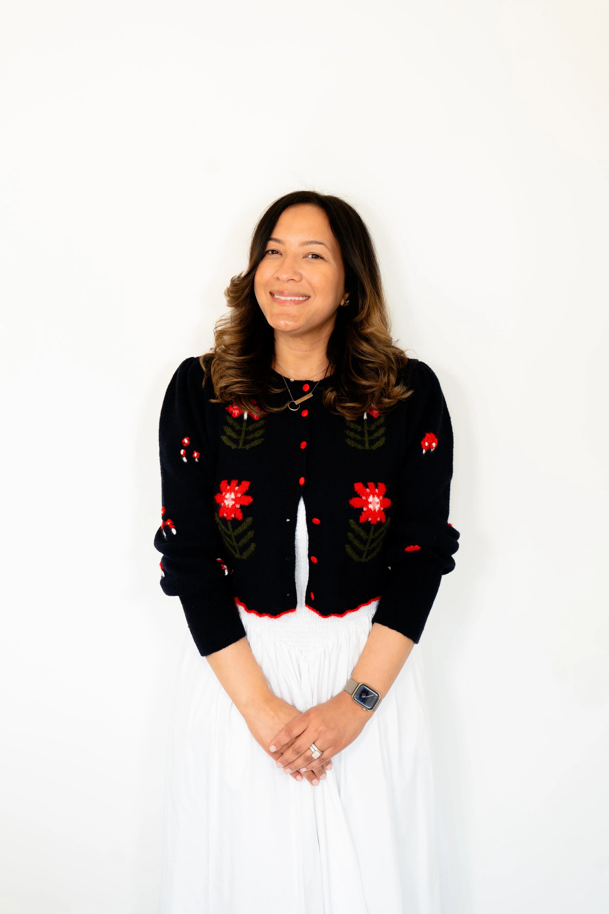 A woman standing against a plain white wall, wearing a black sweater with red and green floral embroidery, a white skirt, and a watch on her left wrist, smiling slightly.