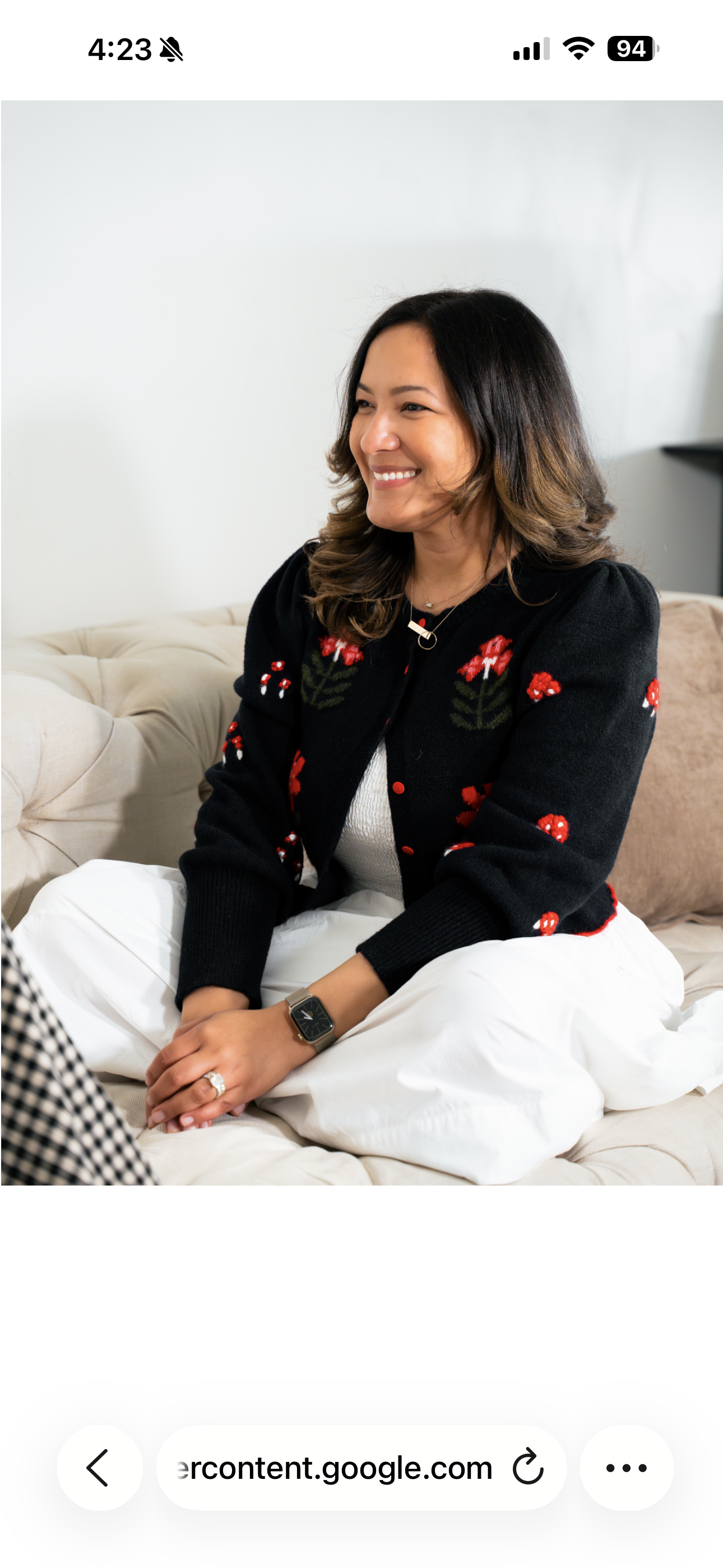 A woman sitting on a beige couch, wearing a black cardigan with red floral embroidery, white pants, smiling and looking to her left.