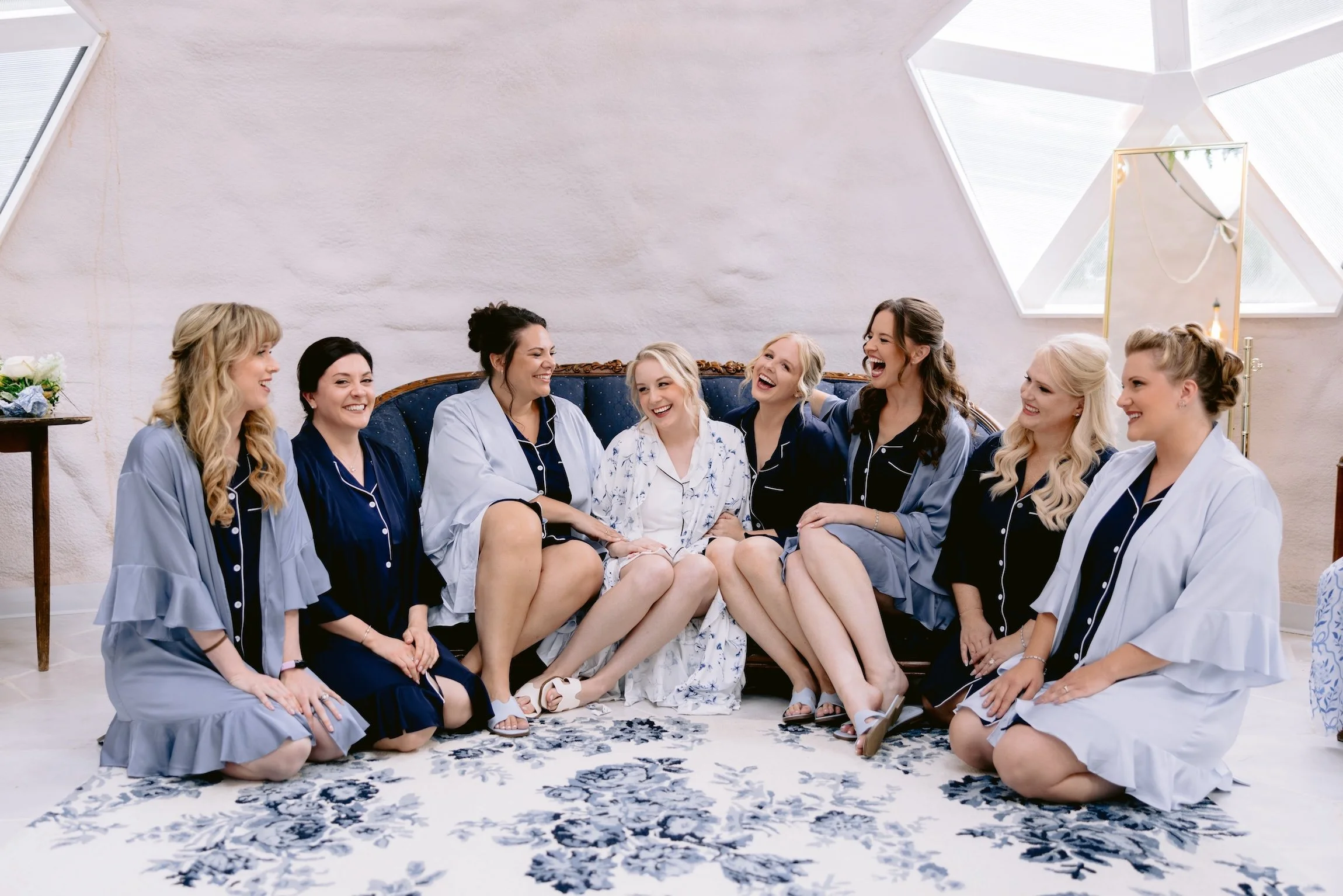 A group of women in pajamas sitting and laughing on a blue patterned rug in a bright room with a high ceiling and hexagonal window.