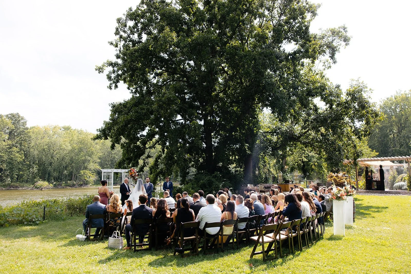 Outdoor wedding ceremony by a river under a large tree with seated guests, officiant, bride, and groom.