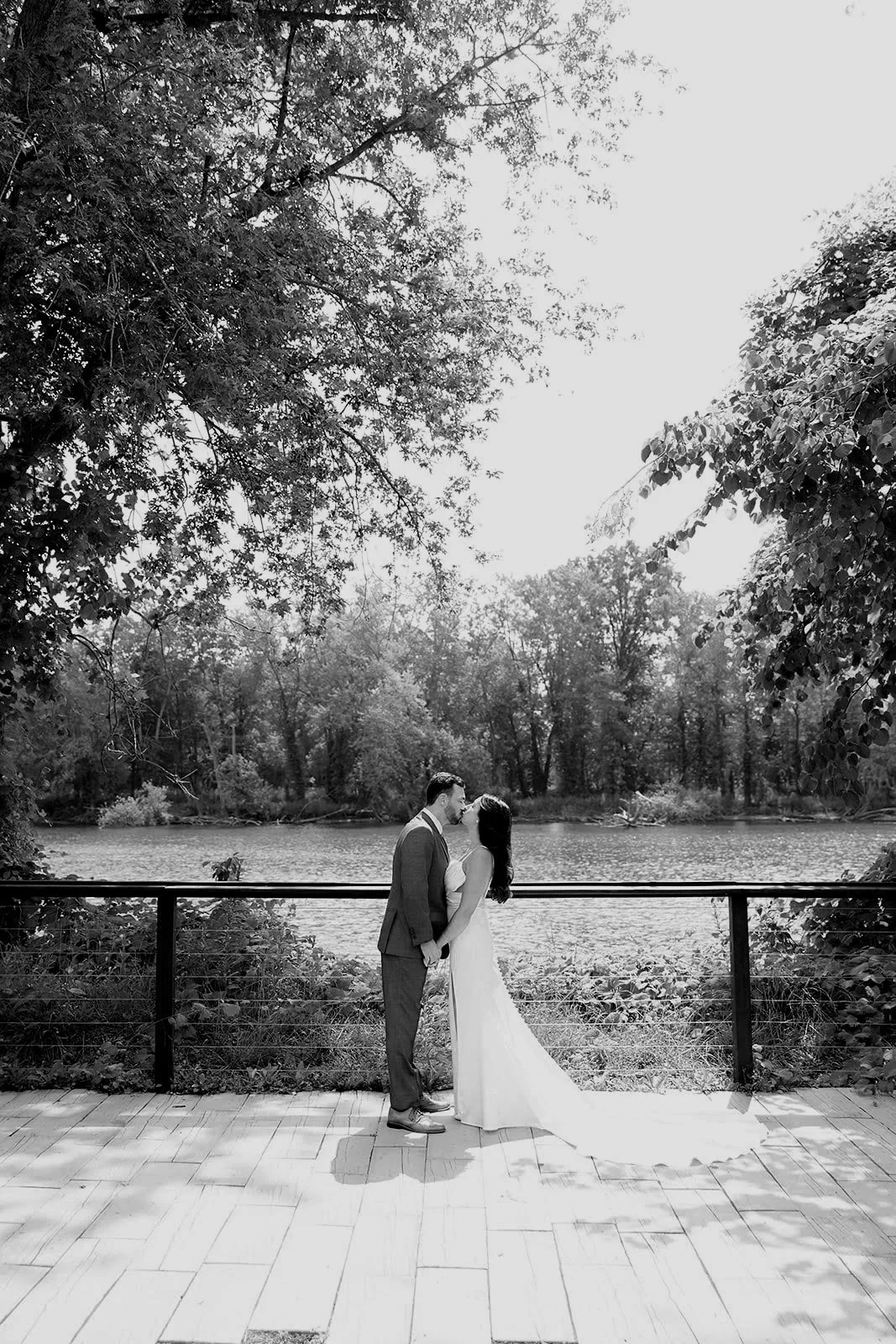A black and white photo of a couple in wedding attire standing close together on a wooden deck near a body of water, holding hands, with trees surrounding them in the background.