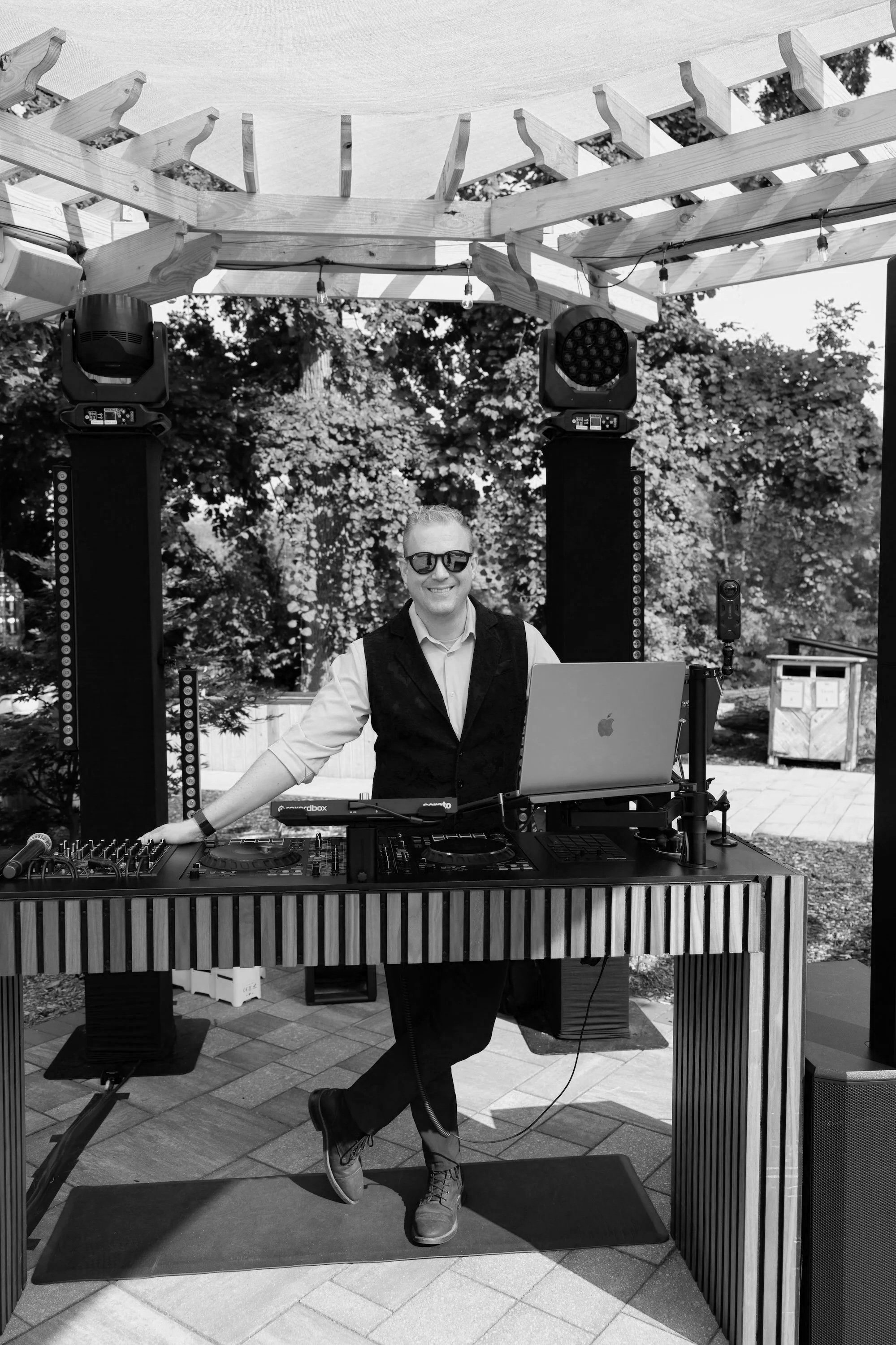 A DJ wearing sunglasses, a white shirt, and a vest standing behind a DJ setup with a laptop and mixing equipment at an outdoor event, with trees and string lights in the background.