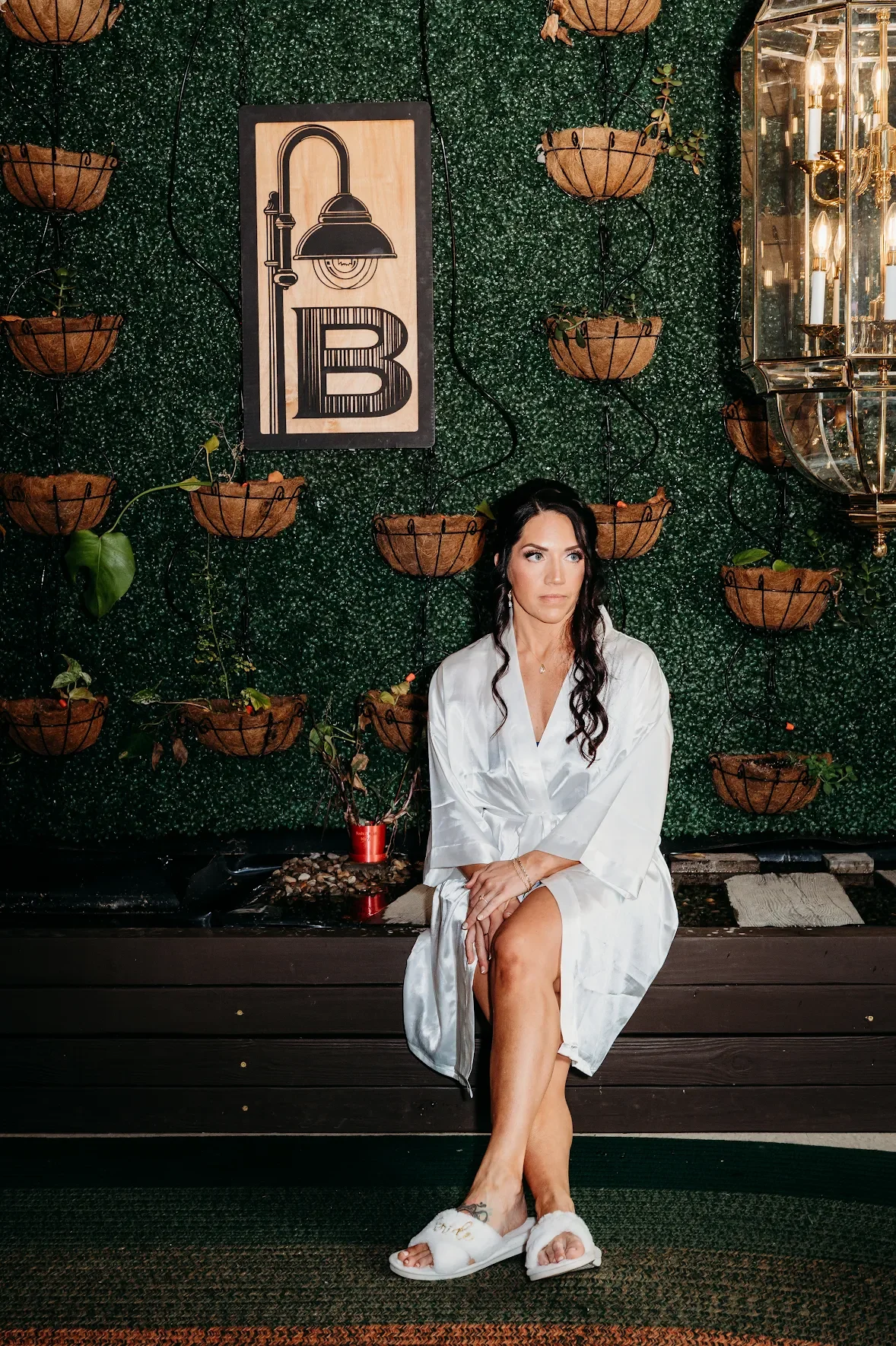A woman sitting on a dark wooden bench in front of a green, leafy wall with hanging brown baskets and a framed art piece featuring a lamp and the letter "B." She is wearing a white satin robe, slippers, and has dark hair with loose curls. She is looking slightly to the side with her hands resting on her lap.