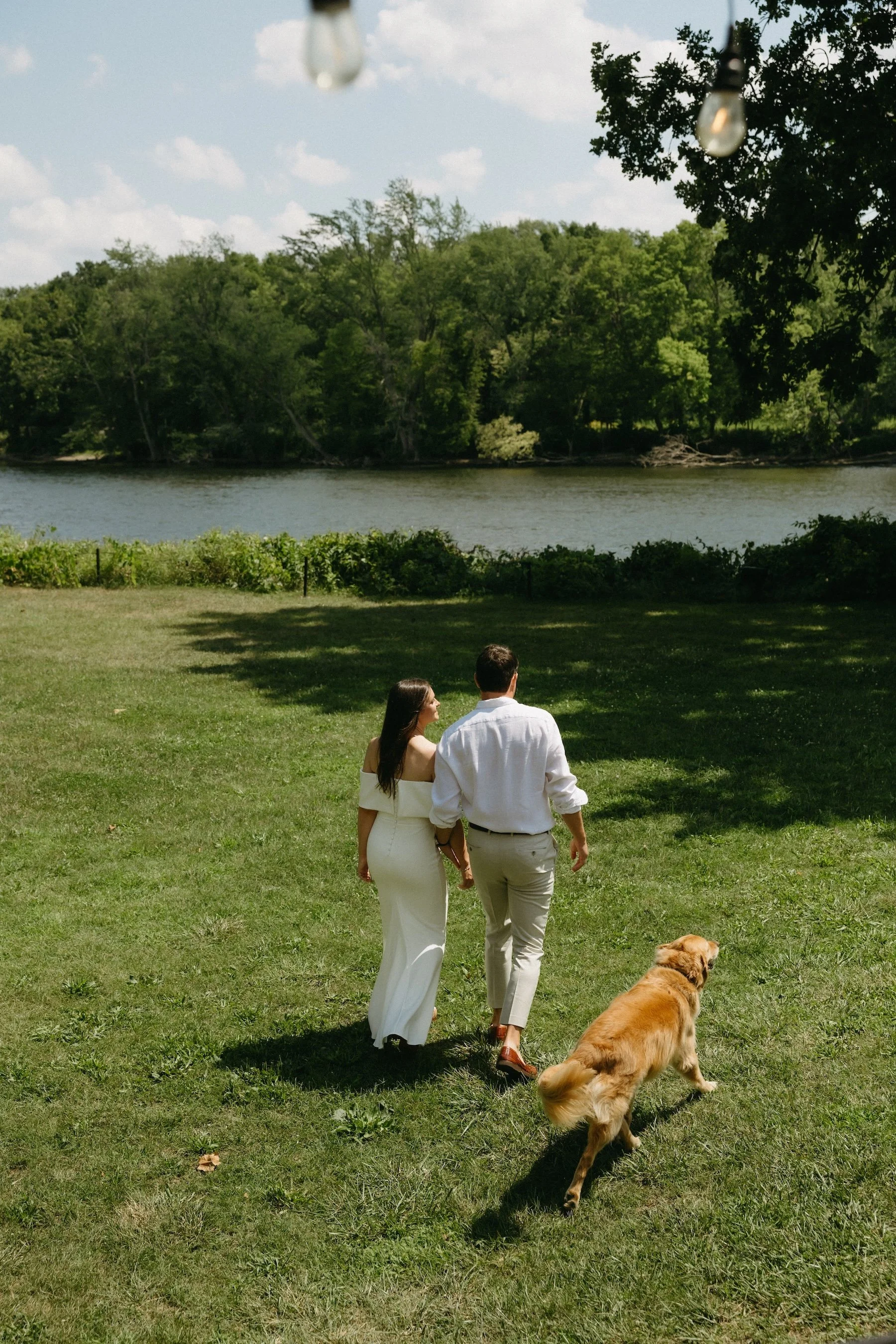 A couple dressed in white walking with a dog in a green park by a lake with lush trees and a blue sky