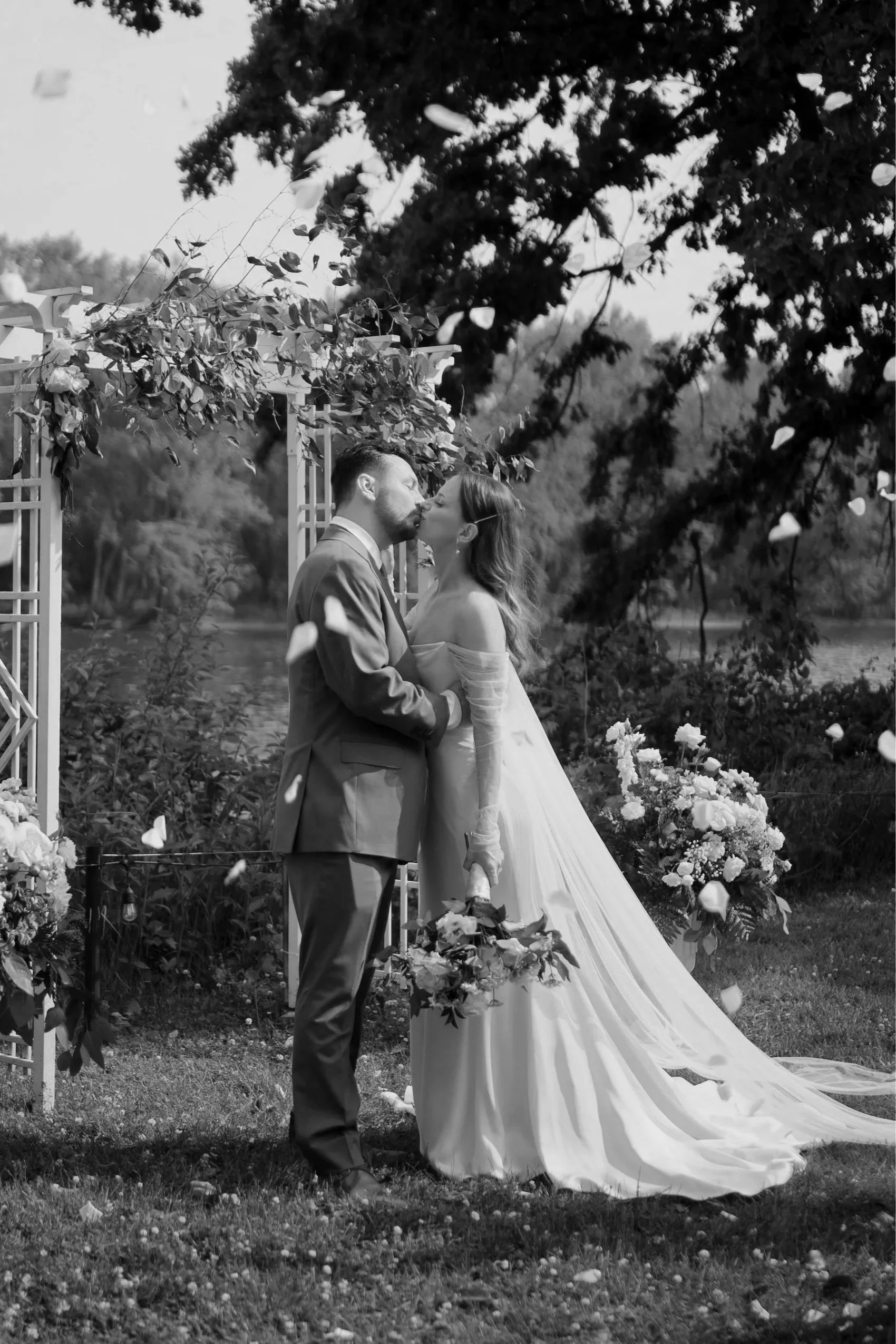 A black and white photo of a bride and groom sharing a kiss during their wedding ceremony outdoors, surrounded by trees and flowers.