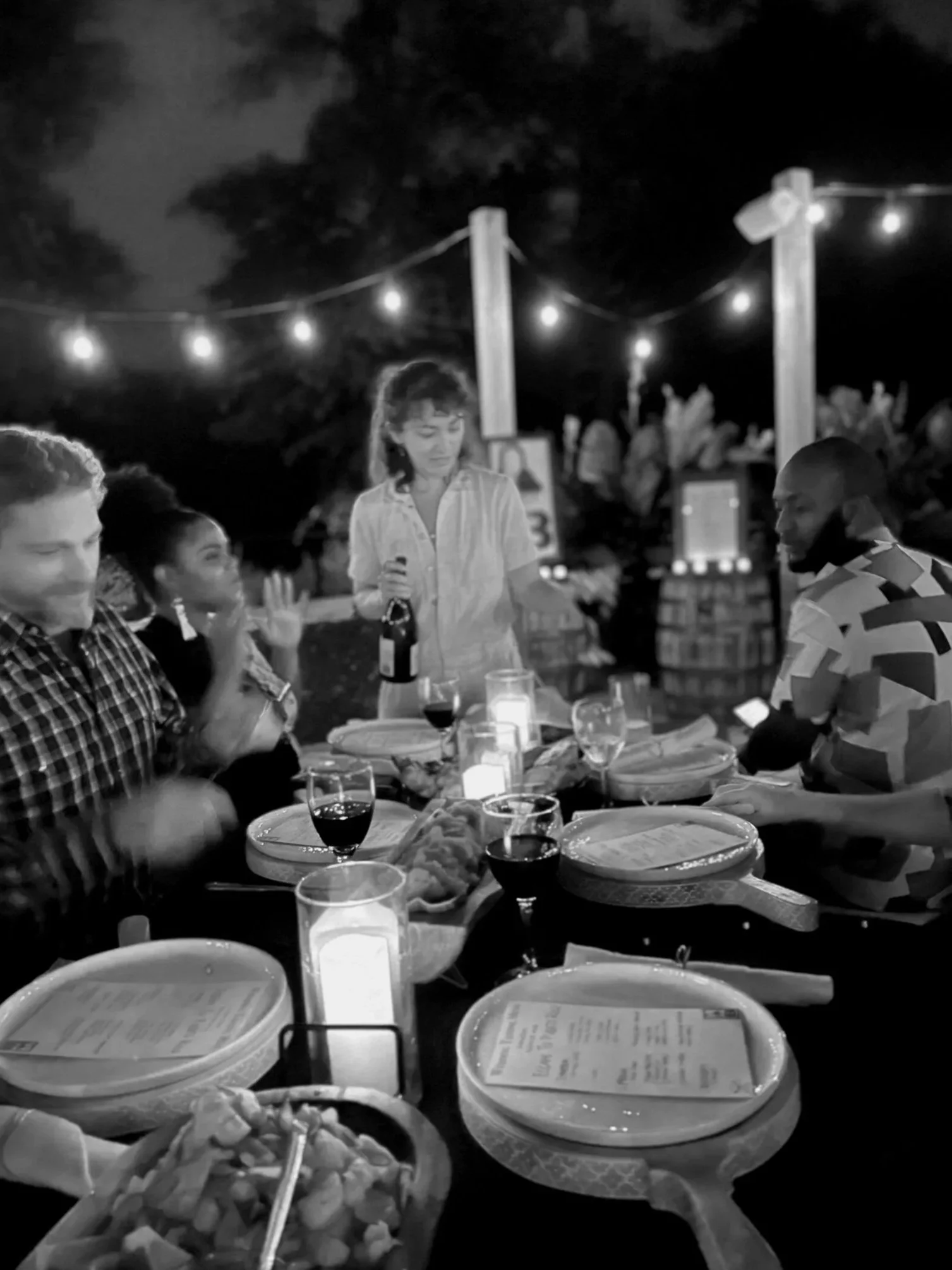 People dining at an outdoor restaurant at night, with string lights overhead, tables set with plates, glasses, and candles.