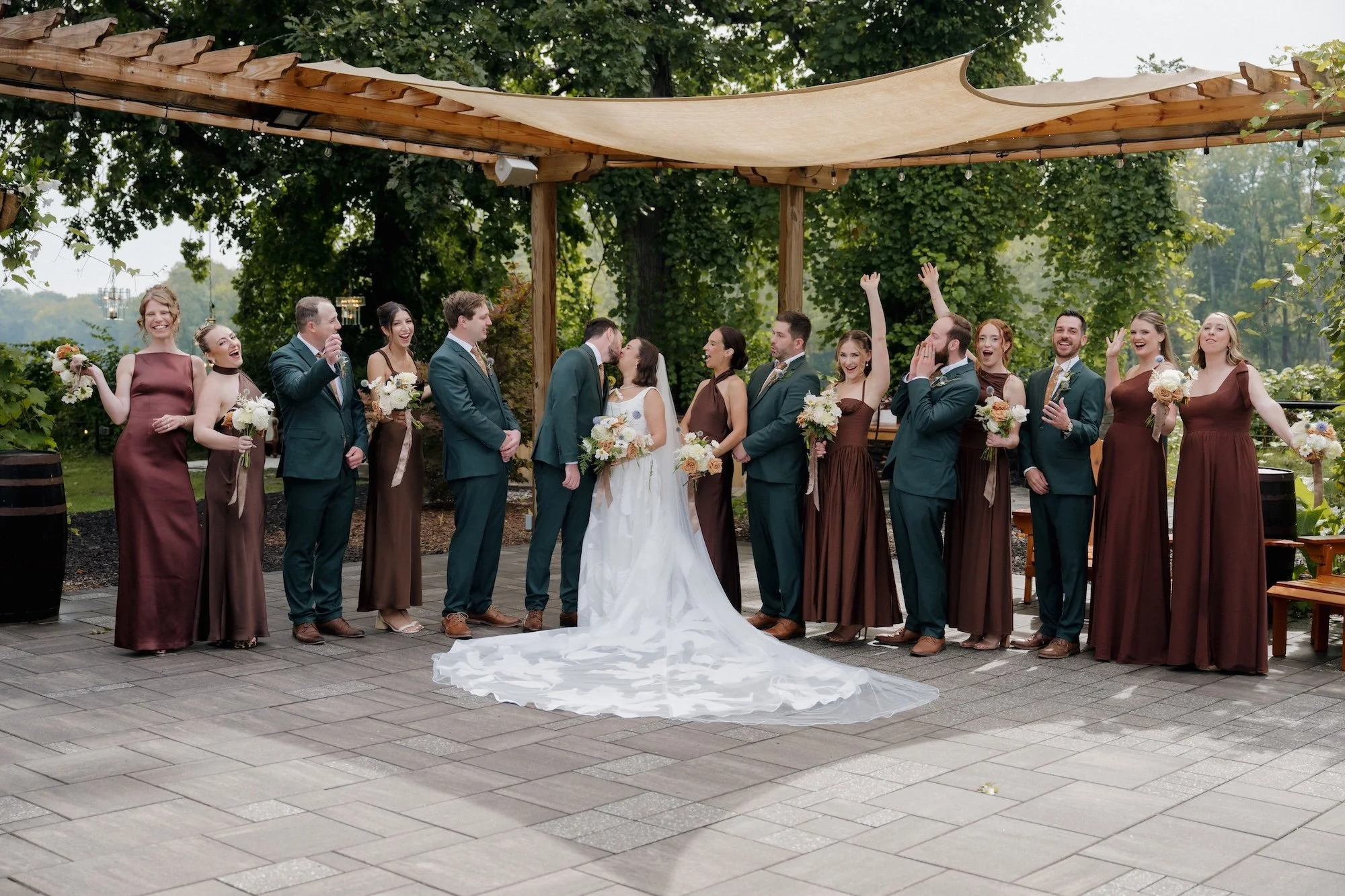 A wedding party stands outdoors under a wooden canopy, with the bride and groom in the center sharing a kiss. The bride wears a white gown with a long train, and the groom wears a dark suit. Bridesmaids in brown dresses and groomsmen in dark suits hold bouquets of flowers and are smiling and celebrating.