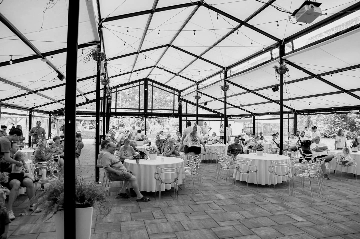 People sitting at round tables in a large, glass-enclosed outdoor event space, with string lights hanging from the ceiling, some people standing, and a few children present.