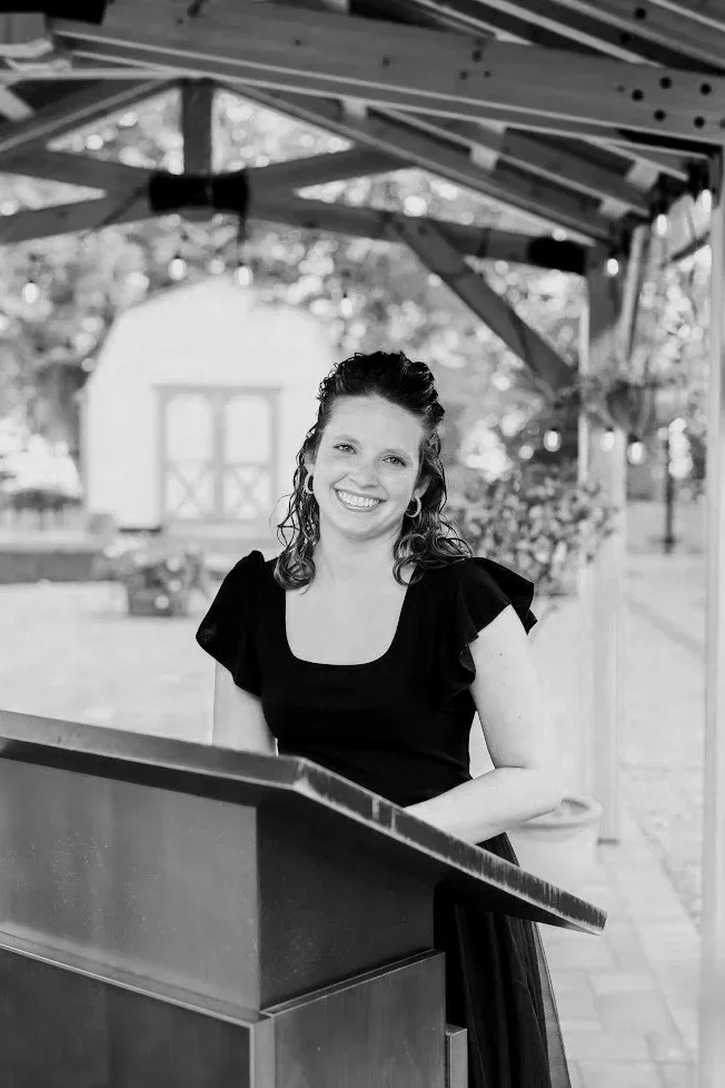 A woman with curly hair smiling, standing behind a piano under a wooden gazebo outdoors, with a garden and shed in the background.