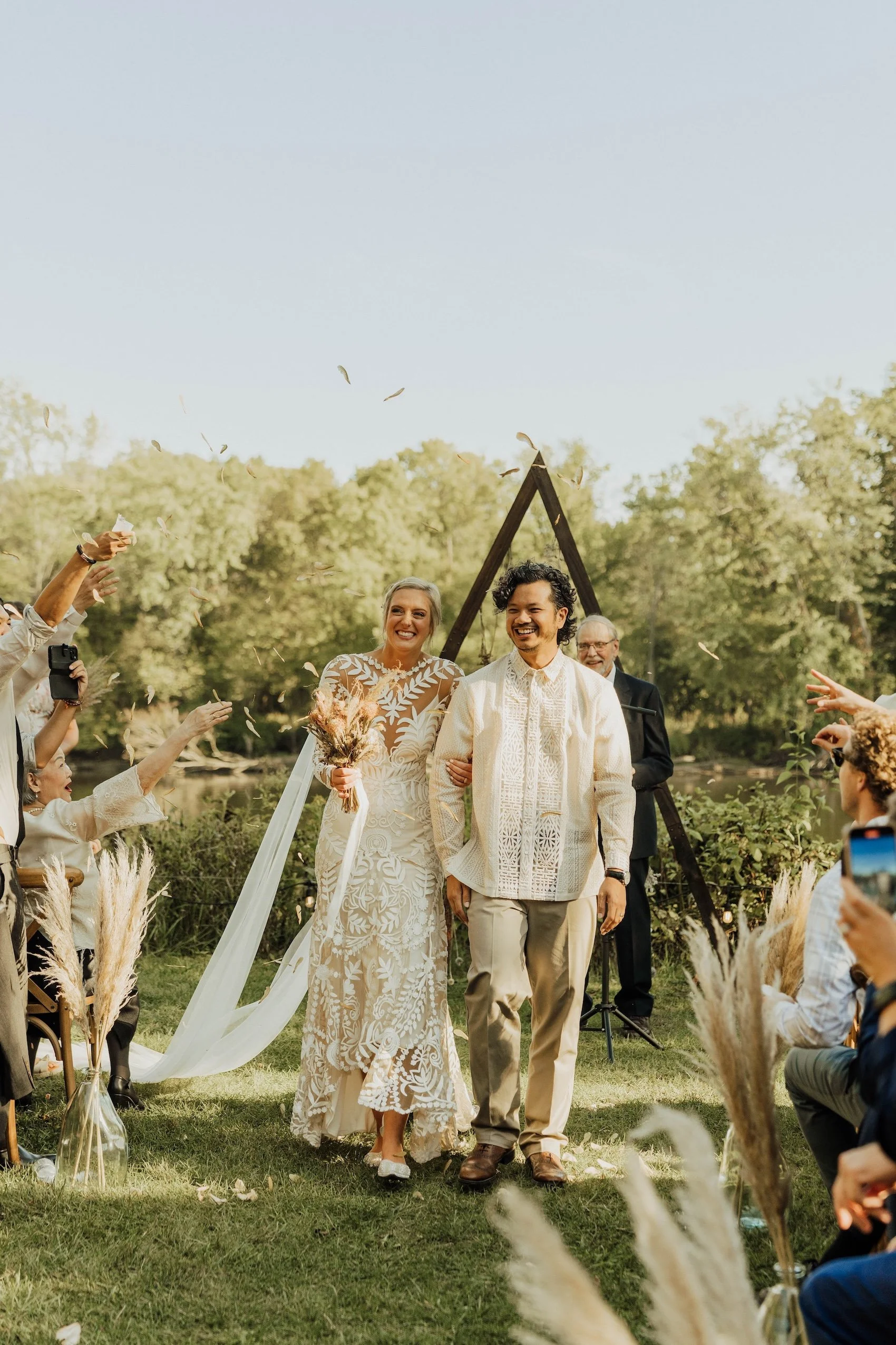 A wedding ceremony outdoors with a bride and groom walking down the aisle, surrounded by guests throwing flower petals, in a natural setting with trees and a clear sky.