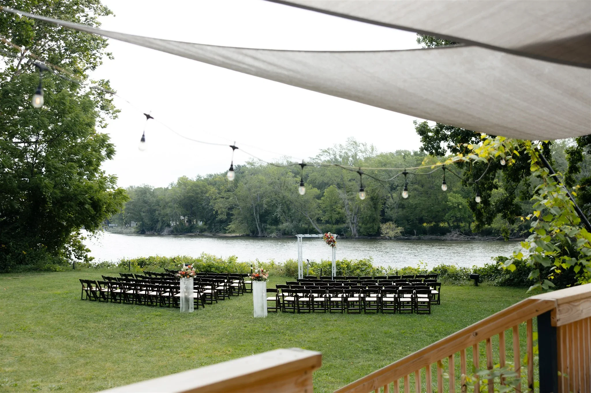 Outdoor wedding setup by a river with chairs and floral arrangements, viewed from a wooden deck with string lights overhead, surrounded by trees.