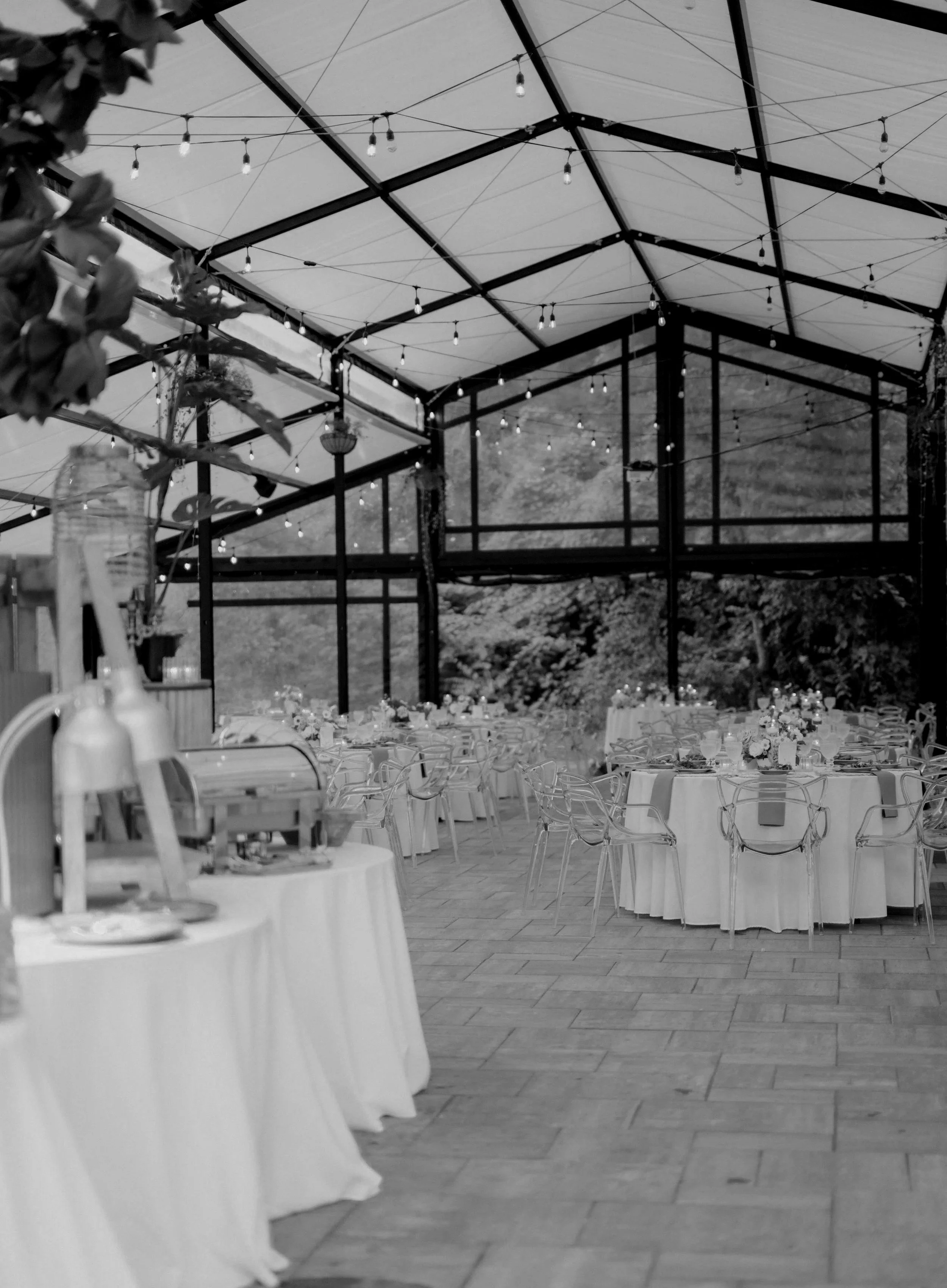Indoor event space with round tables covered in white tablecloths, set for a celebration or banquet, with floral centerpieces and clear chairs. String lights hang from the ceiling, and large windows in the background reveal a mountain landscape.