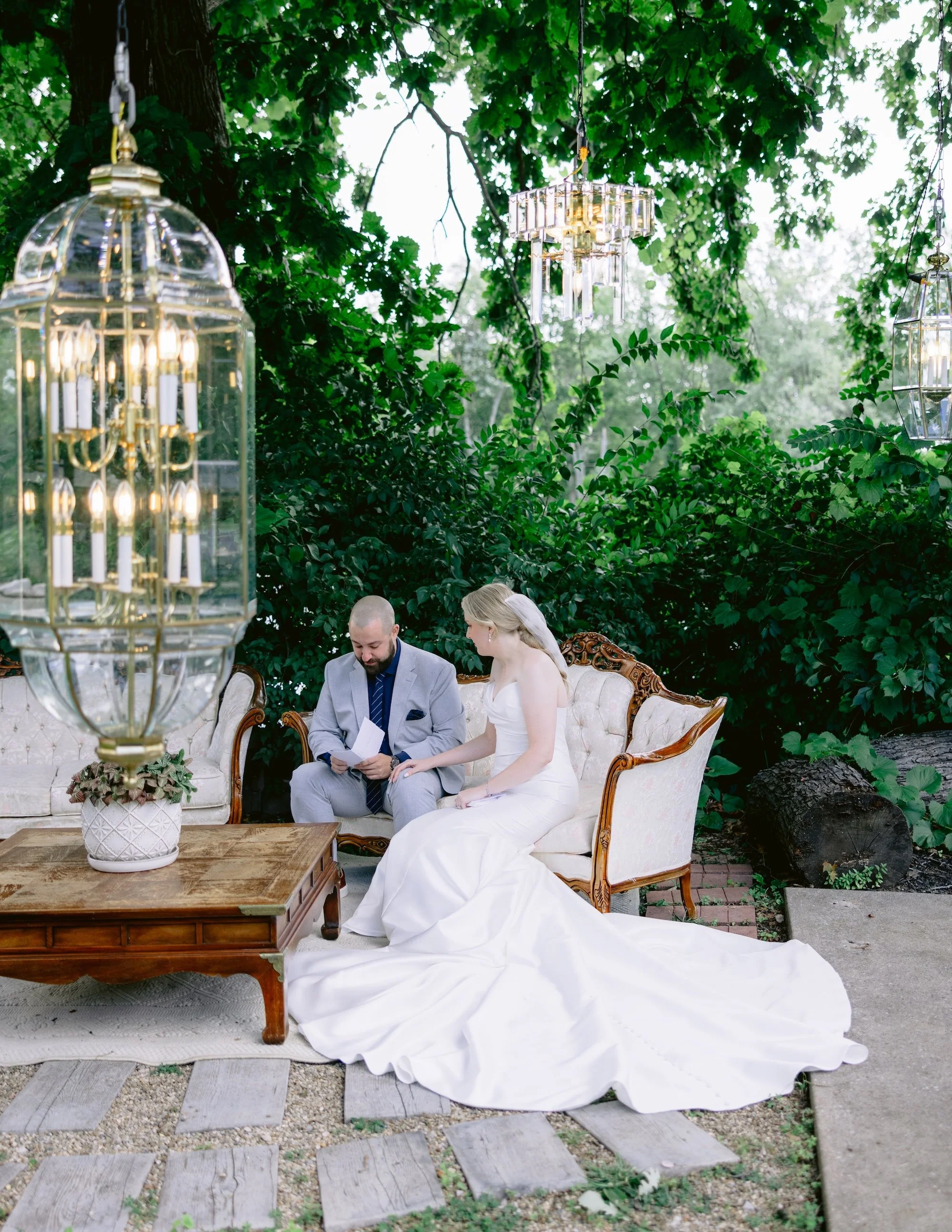 A bride and groom sitting outdoors on an antique sofa during a wedding ceremony, surrounded by lush green foliage and hanging chandeliers.