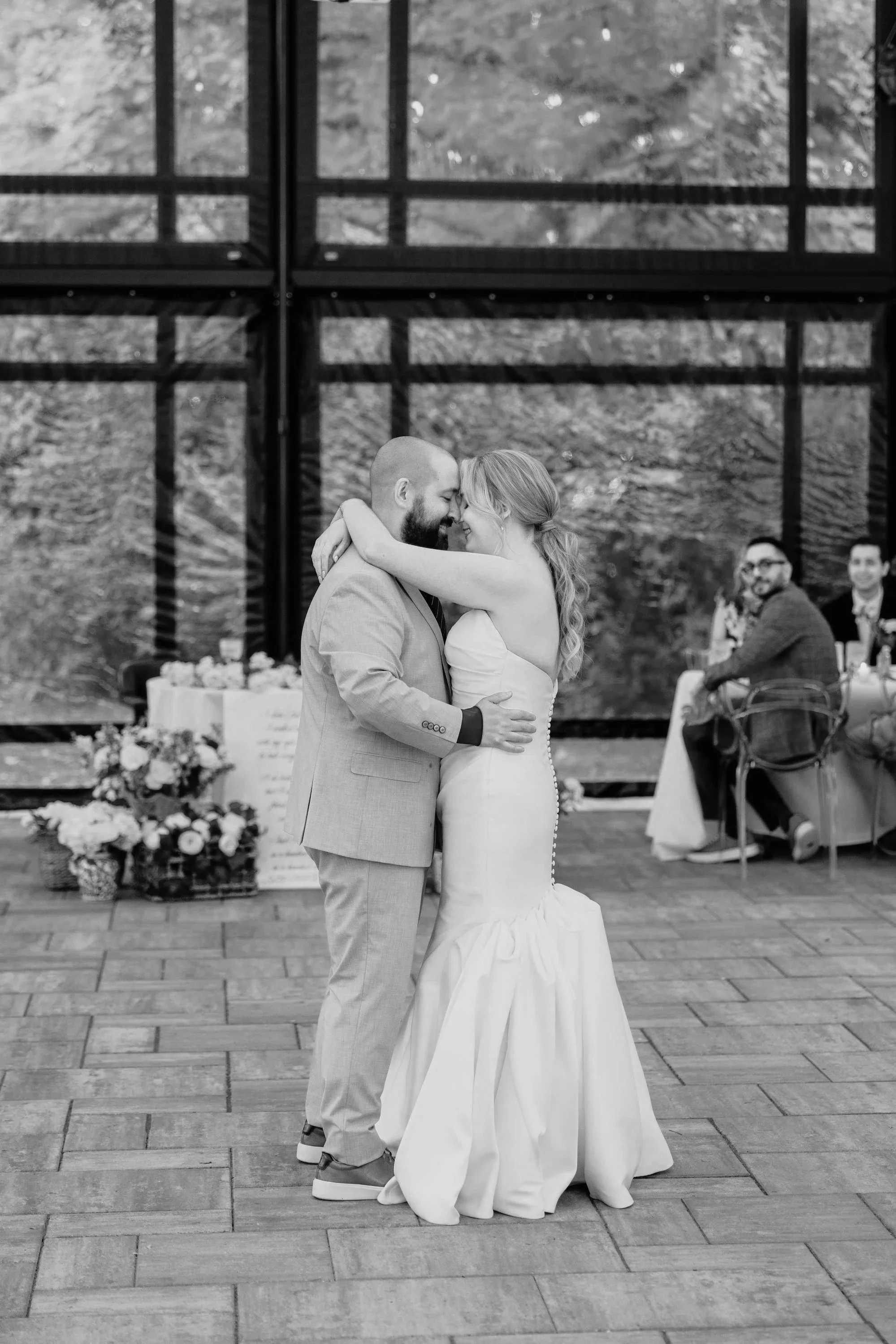A newlywed couple, a man and a woman, are dancing closely during their wedding reception outdoors under a structure with large windows, surrounded by trees and guests seated at tables. The couple is smiling and the woman is wearing a strapless wedding dress, while the man is in a light-colored suit.