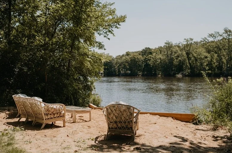 Sandy beach with two wicker chairs and a small table, overlooking a calm river with lush trees on the opposite bank.