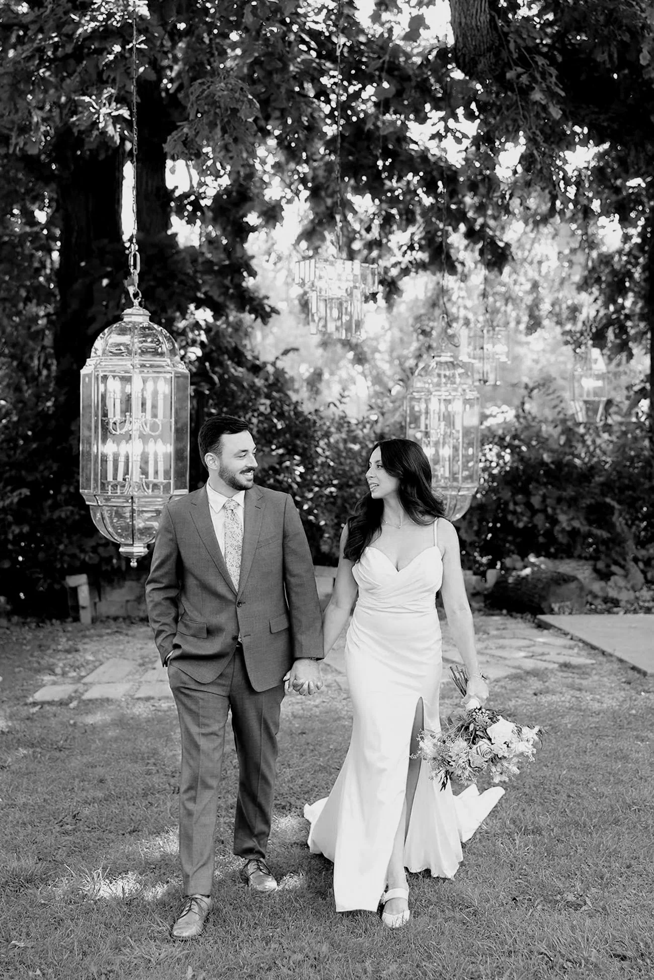 A black and white photograph of a couple walking hand in hand outdoors. The man is dressed in a suit, and the woman is in a white wedding gown holding a bouquet of flowers. They are smiling at each other, surrounded by trees and hanging lanterns.