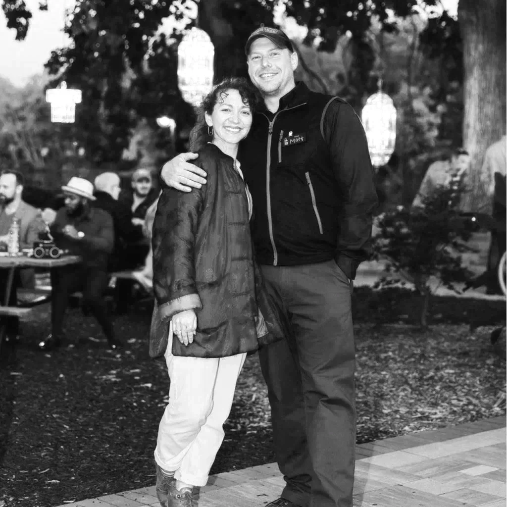 A black-and-white photo of a woman and a man outdoors, smiling, with the woman having her arm around the man's waist. The woman has curly hair and is wearing a loose jacket and pants. The man is taller, wearing a cap, a zip-up jacket, and has a backpack. There are people seated at tables in the background, and hanging lanterns among trees.