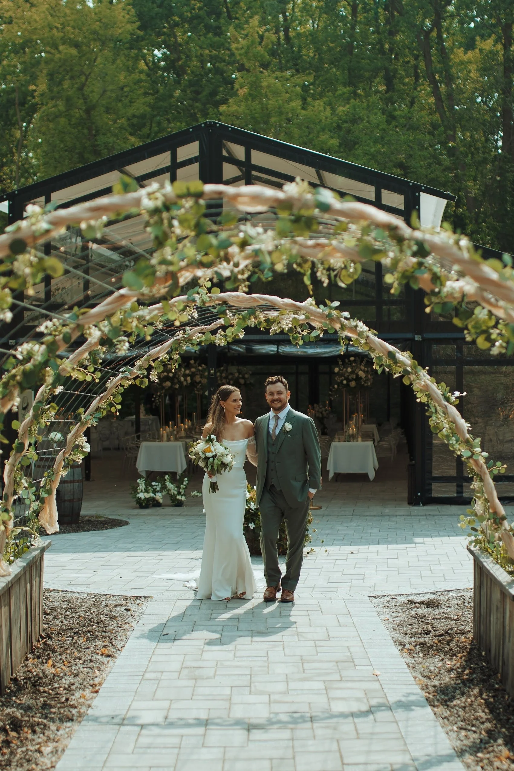 A bride and groom walking under a floral arch at an outdoor wedding venue with a glass building in the background, surrounded by green trees.