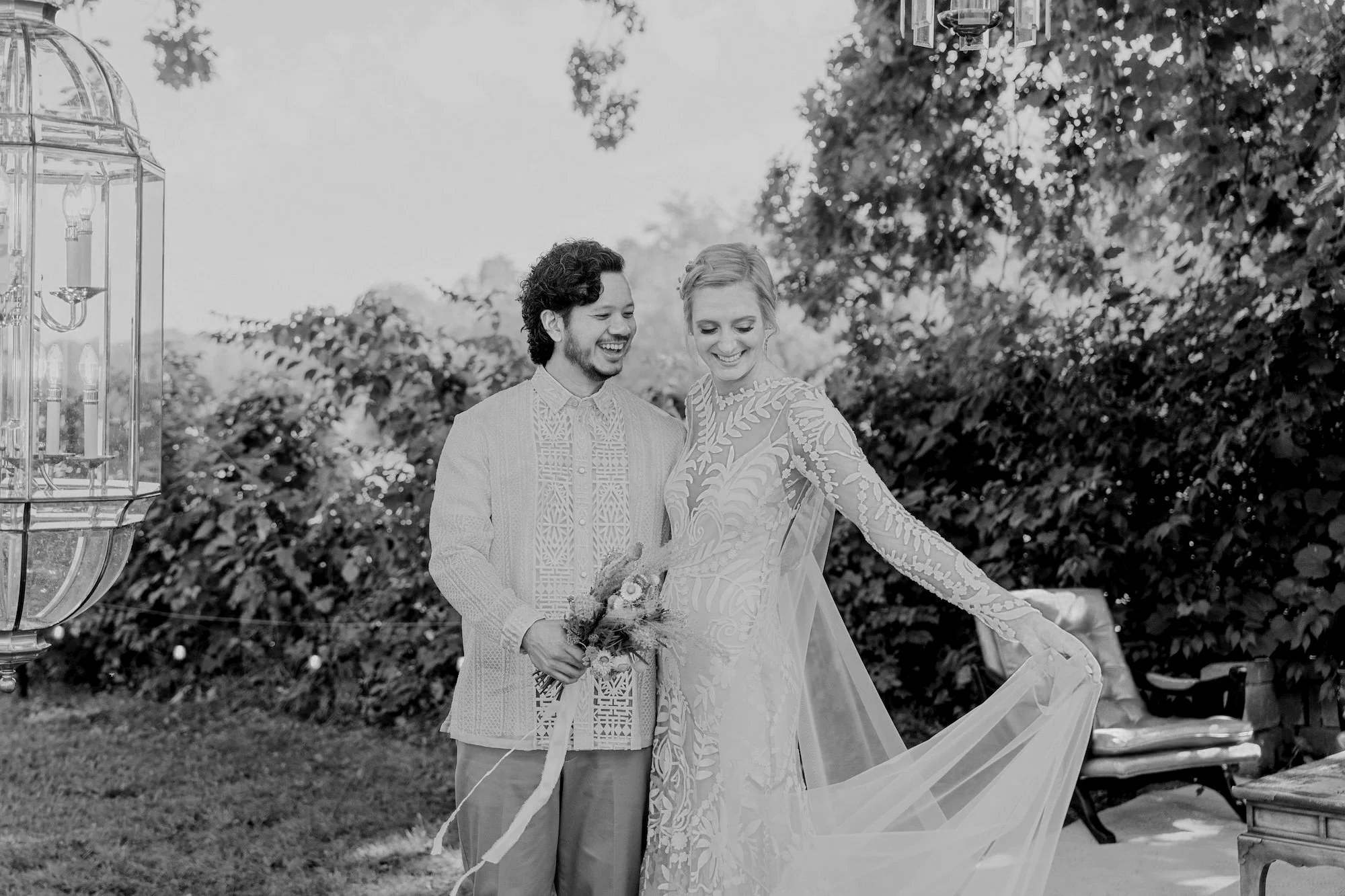 A black and white photo of a happy couple in wedding attire, standing outdoors with trees and bushes in the background. The groom is holding a bouquet and smiling while the bride looks down, smiling, with her hand on a piece of wedding decor.