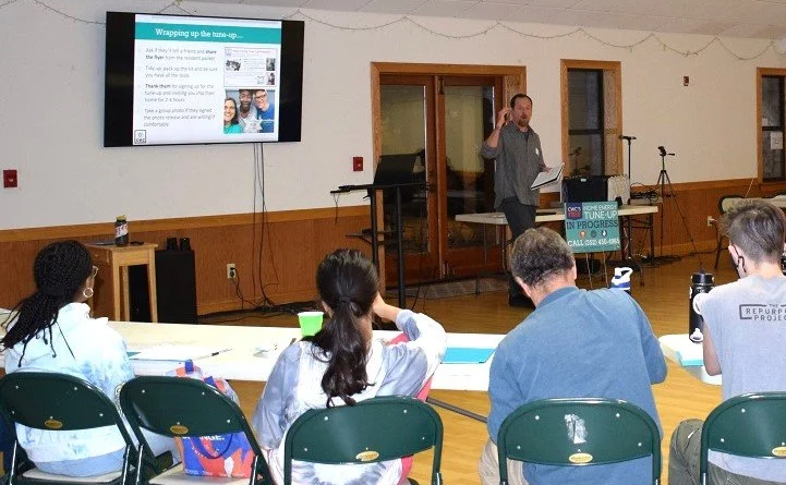 A presentation or workshop taking place in a community center or meeting room. A man stands at the front gesturing as he speaks, with a large screen displaying information behind him. Four attendees sit at a table facing the presenter, some with notebooks and beverages in front of them.