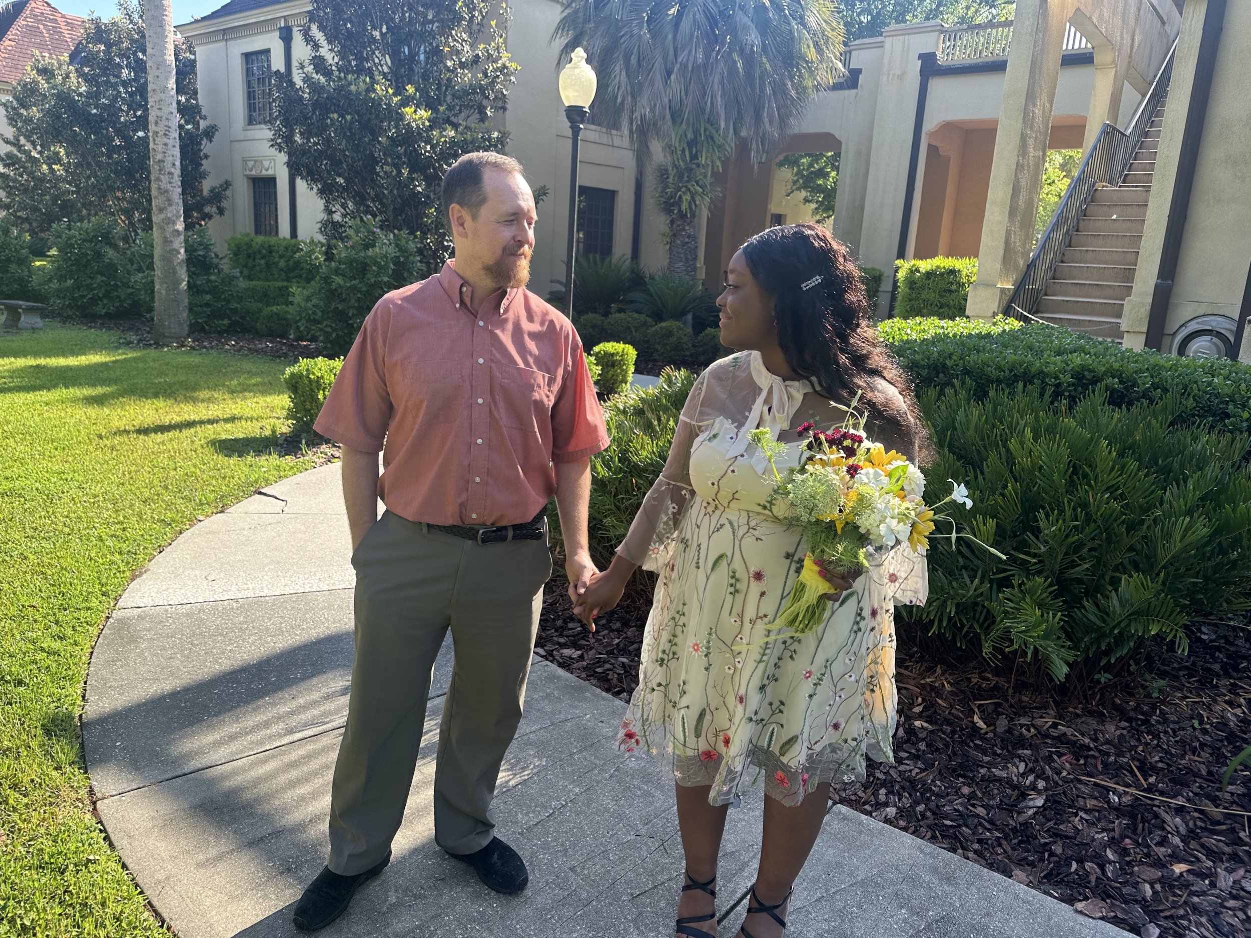 A man and woman holding hands and looking at each other outdoors in a garden with green bushes and trees, a building in the background, and a woman holding a bouquet of flowers.