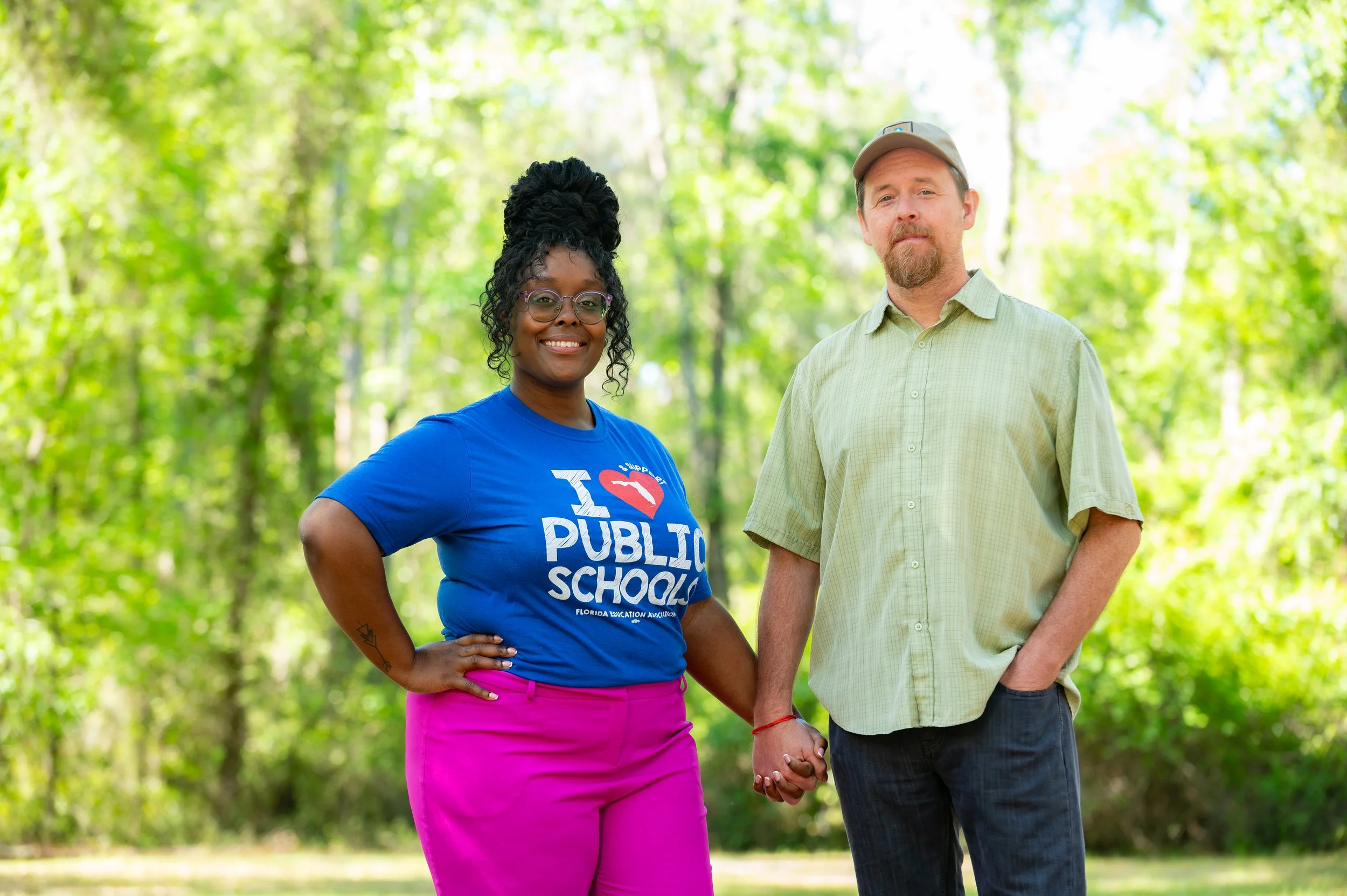 A smiling woman and a man holding hands in a green outdoor setting with trees and sunlight. The woman is wearing a blue T-shirt and pink pants, and the man is wearing a light green shirt and a beige cap.