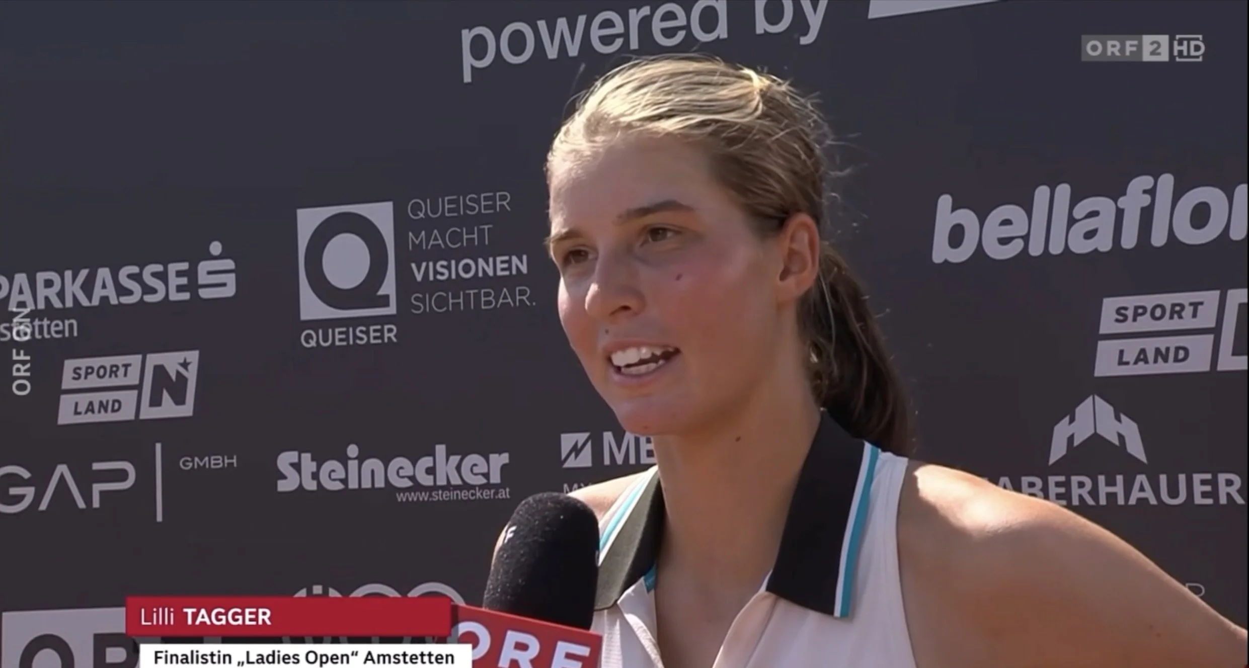 Lilli Tagger bei einem ORF-Interview im Rahmen der Live-Übertragung des Finales 2025. Hinter ihr eine Dunkle Wand mit Sponsorenlogos. Im TV-Insert steht 'Lilli Tagger, Finalistin Ladies Open Amstetten' auf Deutsch.