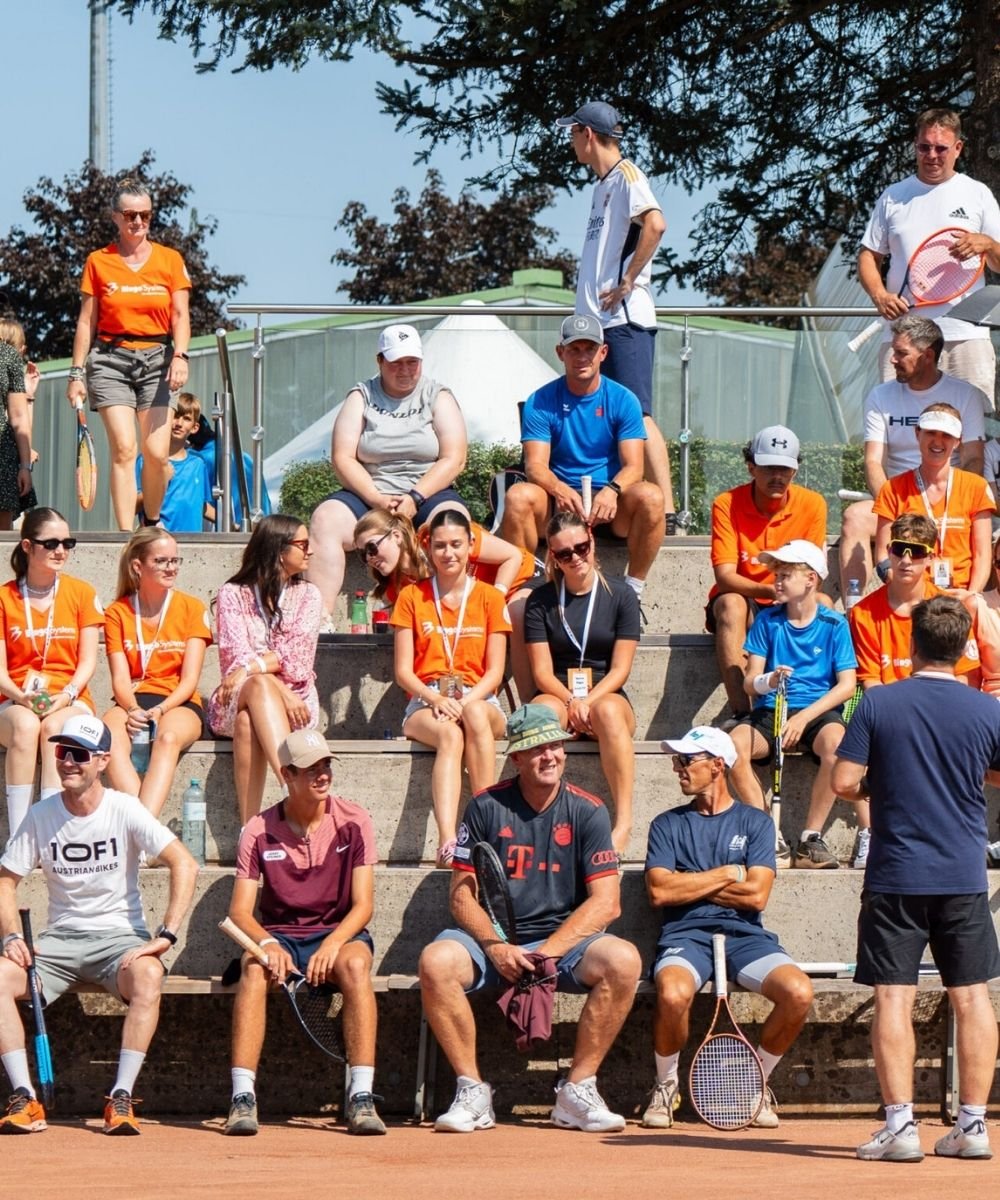 Menschen sitzen und stehen auf einer Tribüne bei einem Tennisevent, einige tragen Tennisausrüstung und Sonnenhüte, im Hintergrund Bäume und ein blauer Himmel.