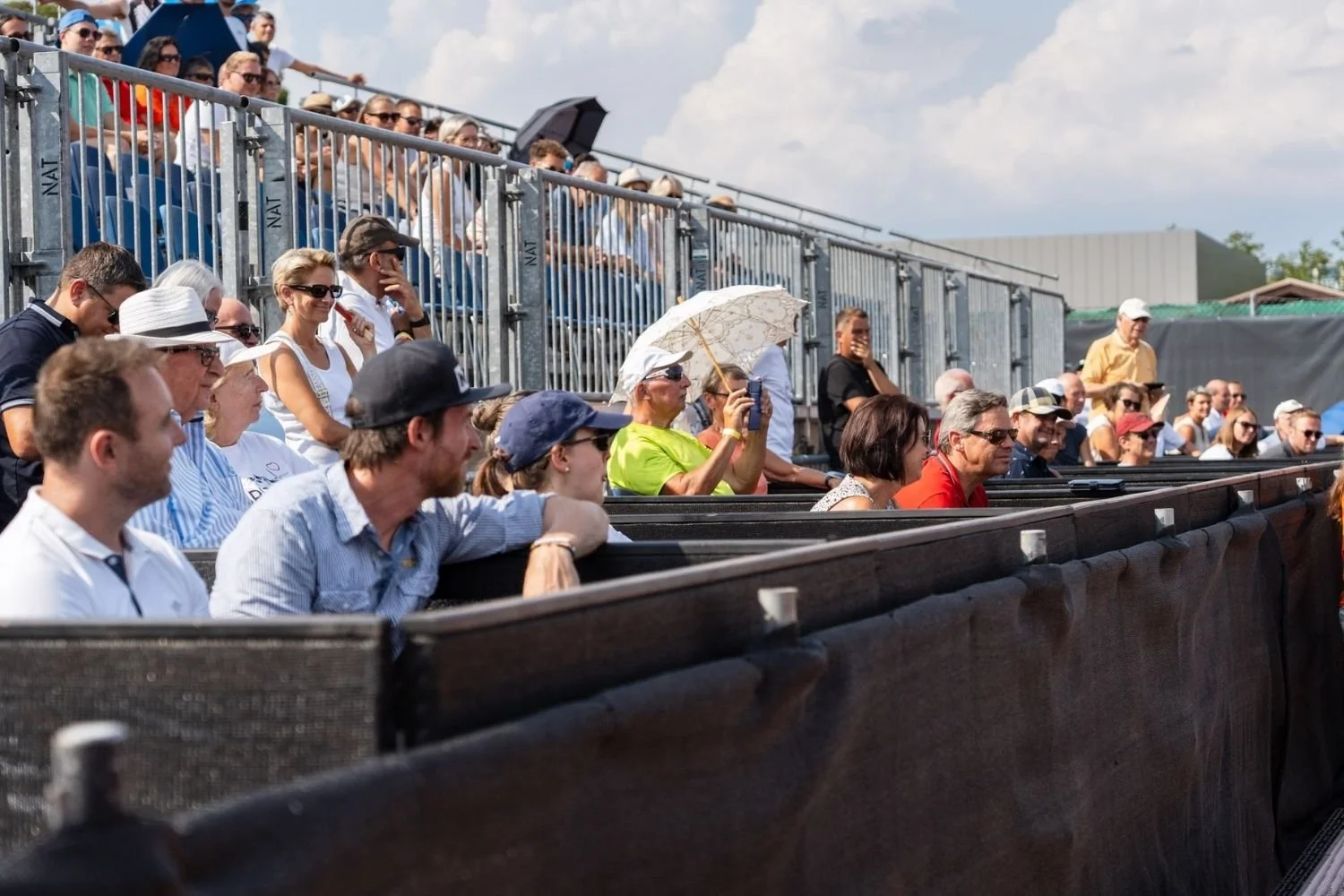 Menschen sitzen in den VIP-Boxen der LADIES OPEN Amstetten, einige mit Sonnenbrillen und Hüten, eine Fan fotografiert mit einem Smartphone.