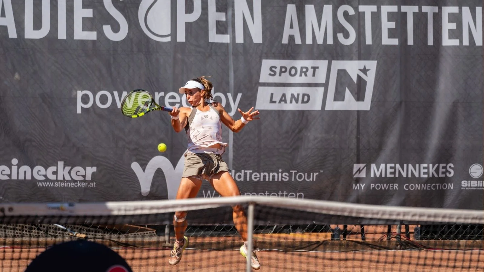 Eine Frau im weißen Sportoutfit und einer White Cap spielt Tennis auf einem roten Sandplatz. Sie bereitet sich vor, den Tennisball mit ihrem Schläger zu treffen, während das Netz im Vordergrund sichtbar ist.