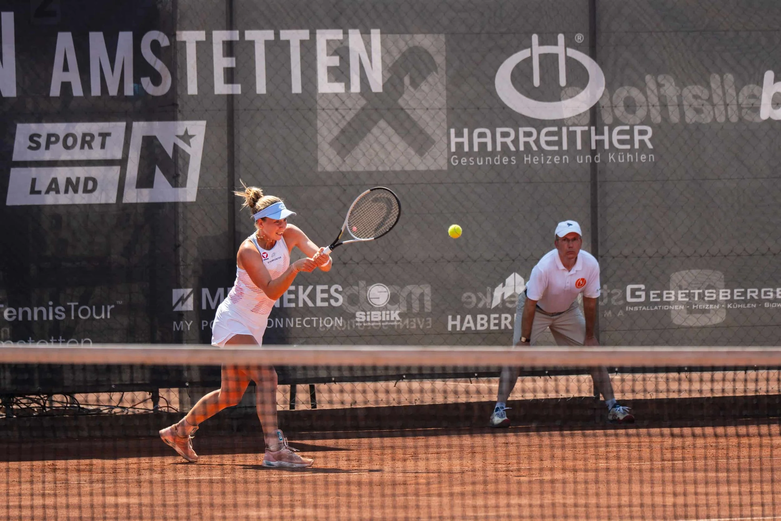 Junge Frau beim Tennis, die den Ball schlägt, während ein Mann zuschaut, auf einem roten Tennisplatz in Deutschland.