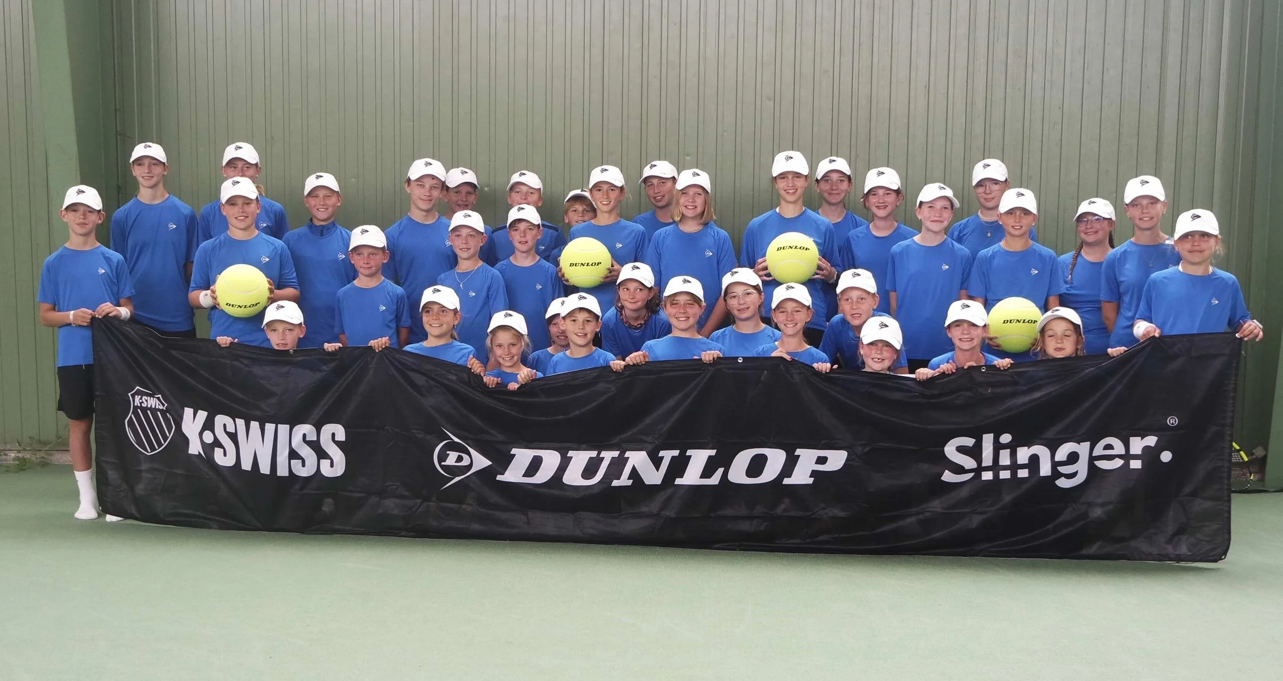 Gruppenbild eines Kinder-Tennis-Teams in blauen Trikots und weißen Caps, die zusammen auf einem Tennisplatz stehen, einige halten Tennisbälle, im Vordergrund eine schwarze Fahne mit Markenlogos.