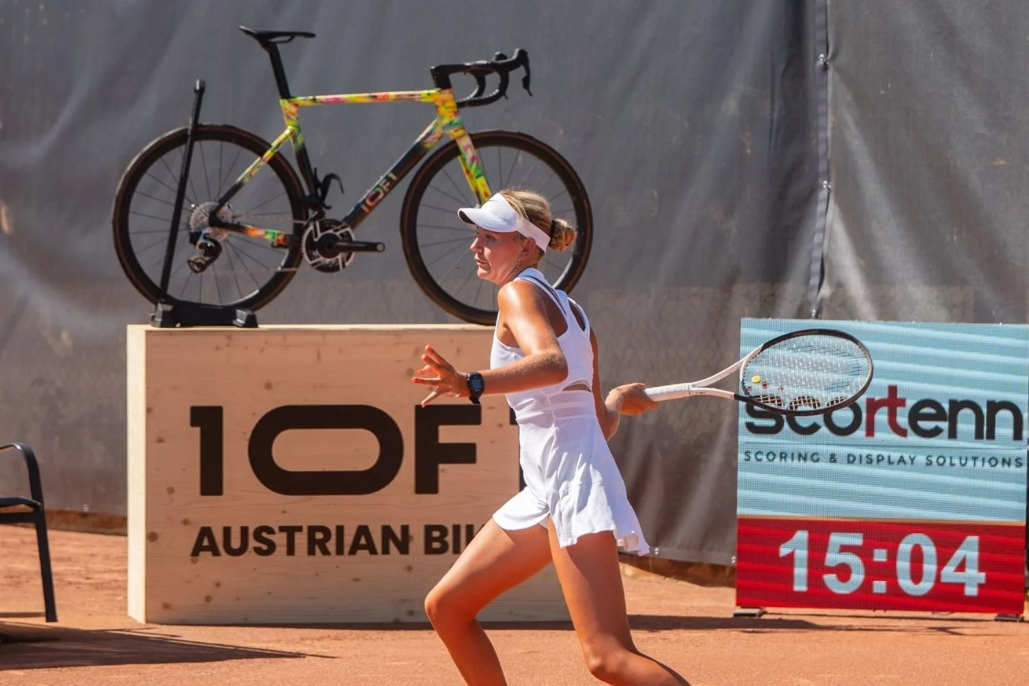 Eine Tennisspielerin läuft auf einem Sandplatz während eines Spiels. Im Hintergrund ist ein Fahrrad auf einem Podest neben einem Schild mit der Aufschrift '10F1- Austrian Bikes' und einem digitalen Timer, der 15:04 anzeigt.