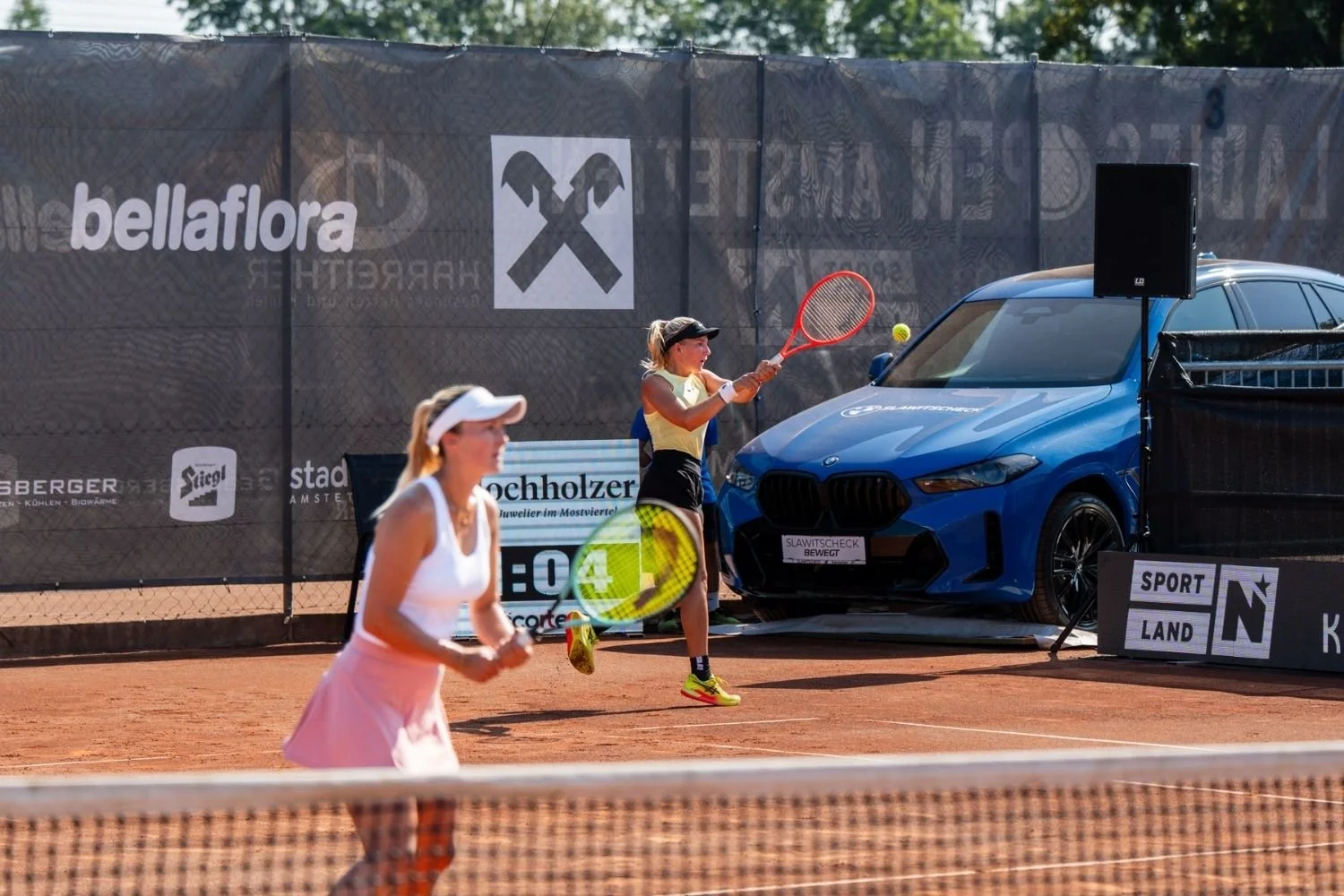 Zwei junge Frauen spielen Tennis auf einem roten Sandplatz, während neben dem Spielfeld ein blauer BMW geparkt ist und ein Lautsprecher steht. Die Frau im Hintergrund schlägt den Tennisball, während die andere im Vordergrund läuft.
