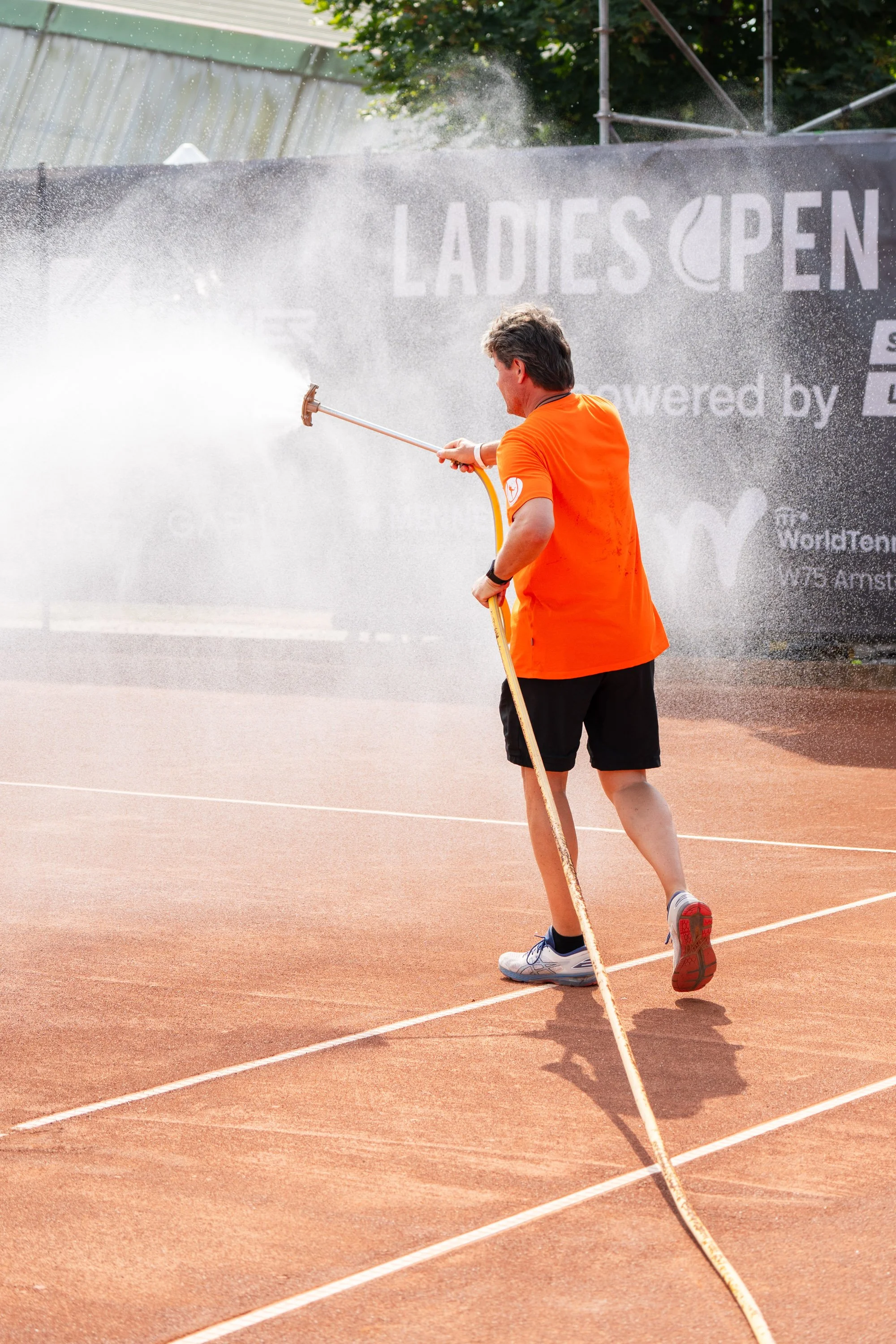 Ein Mann in orangefarbenem T-Shirt und schwarzen Shorts, der den Tennisplatz in Amstetten spritzt.