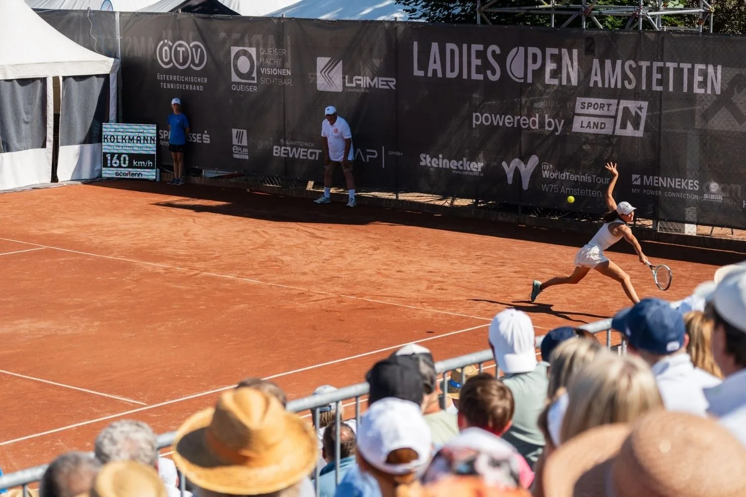 Eine Tennisspielerin auf einem roten Sandplatz während eines Turniers, die sich aus der Position begibt, während sie den Ball im Spiel hat, mit einem Publikum im Vordergrund und einem Banner im Hintergrund.